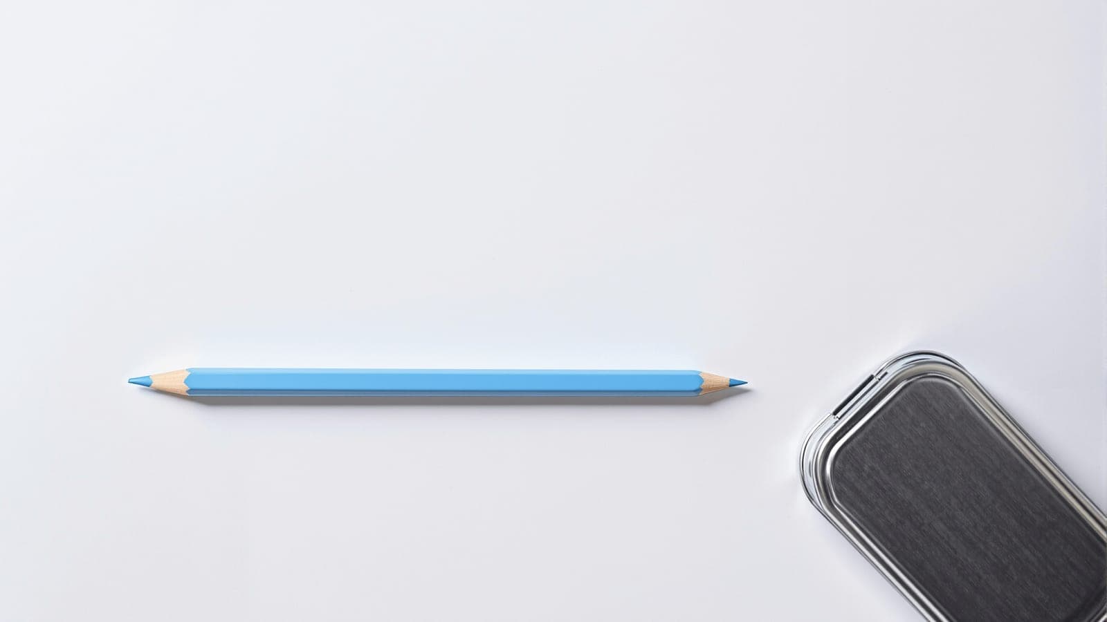 A single coloured pencil lying alone on white paper beside a closed metal pencil tin, the rest of the colours shut away inside