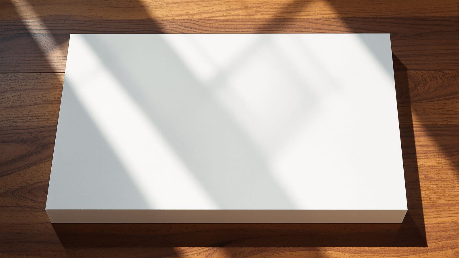 A plain rectangular foam base sheet lying alone on a wooden desk in raking light, its long axis catching the sun