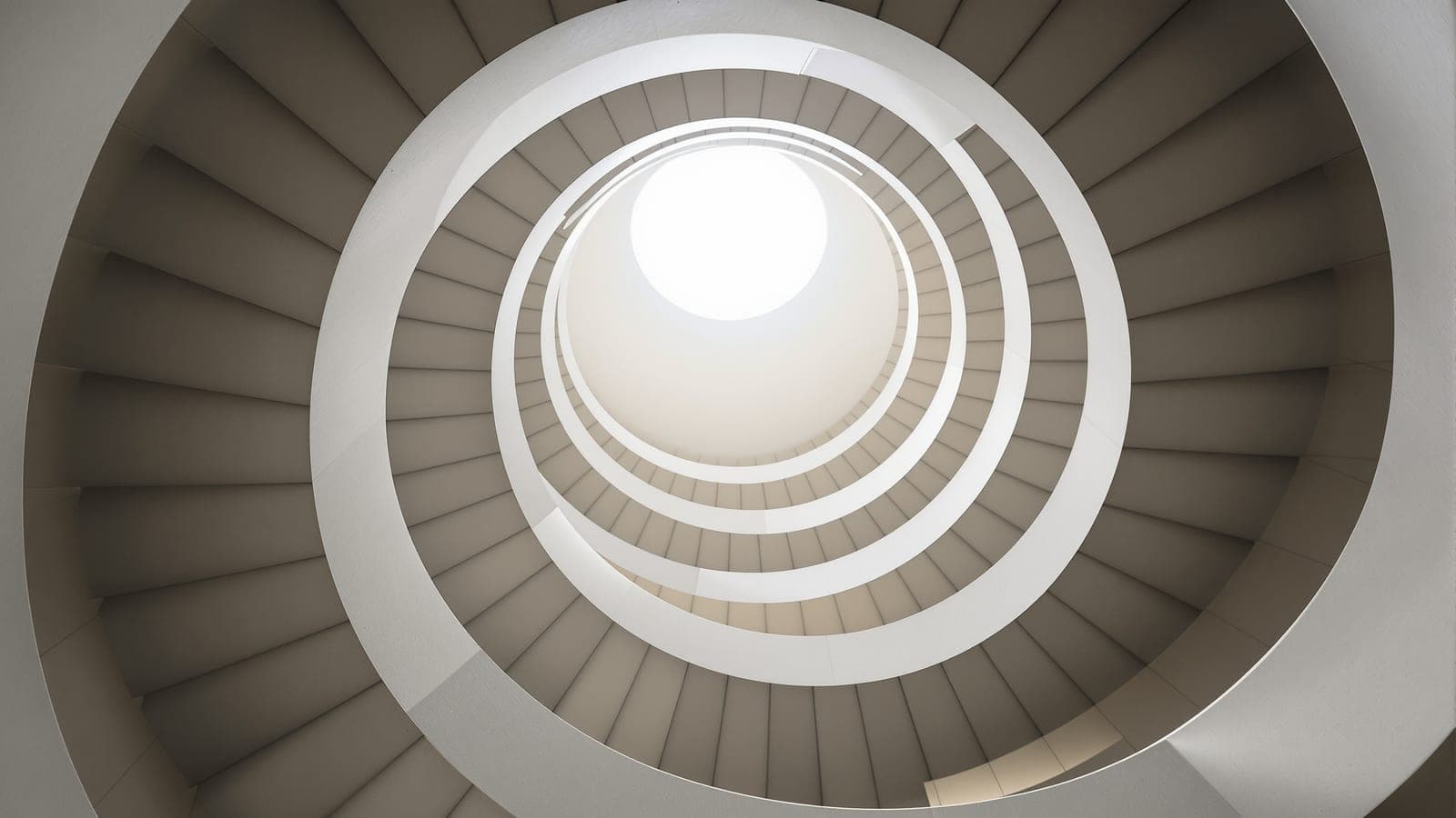 A spiral staircase seen looking straight up from below, its steps repeating in a strict rule toward a bright opening at the centre