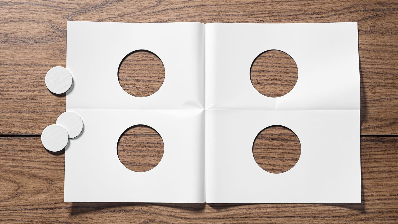 An unfolded sheet of paper on a wooden desk showing two crease lines and four punched holes arranged symmetrically about them, the punched discs lying alongside