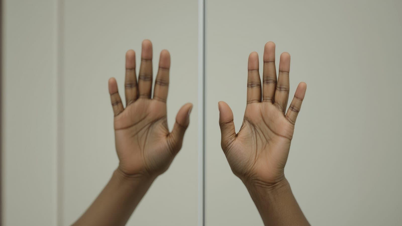 A brown-skinned hand raised palm-out at a mirror beside its own reflection, the two forming a mirrored pair that cannot be rotated onto each other