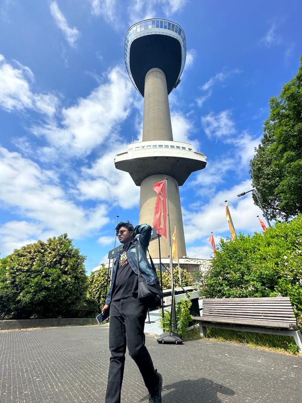 Amogh standing at the foot of the Euromast in Rotterdam, the concrete tower and its crow's-nest platform rising into a blue sky behind him