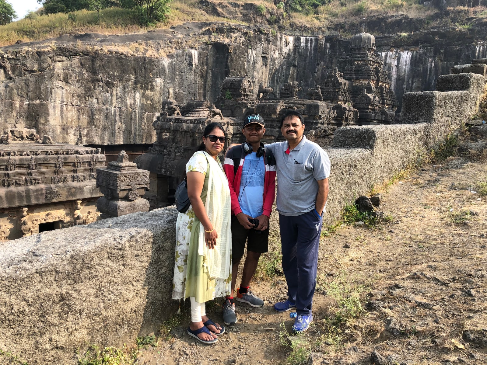 Amogh with his mother and father in the sunlit courtyard of the Kailasa temple at Ellora, a camera in his hand, the monolithic rock-cut shrines and the basalt escarpment of the caves rising behind them