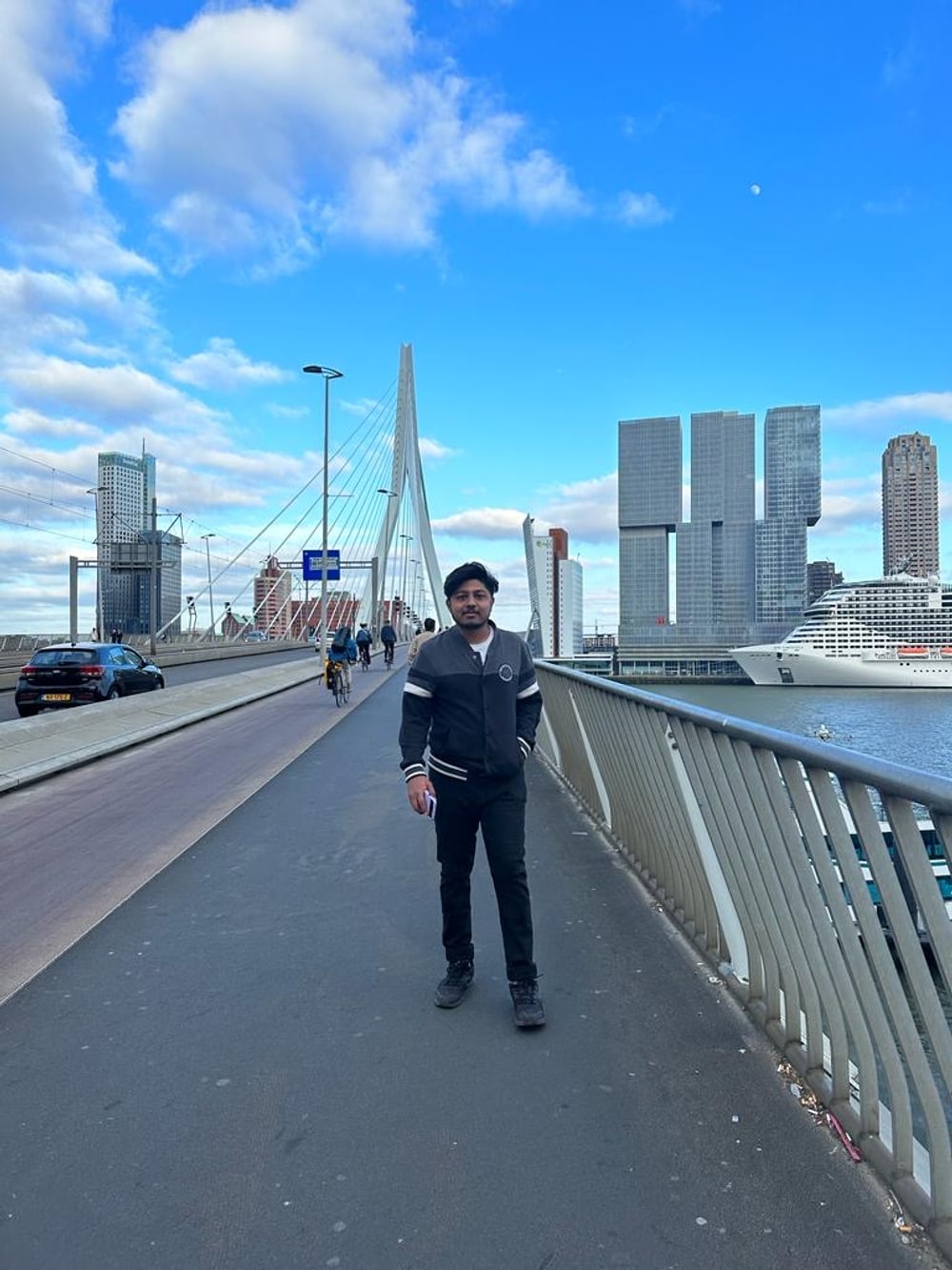 Amogh standing on the Erasmus Bridge in Rotterdam, the towers of the Kop van Zuid skyline rising behind him across the water
