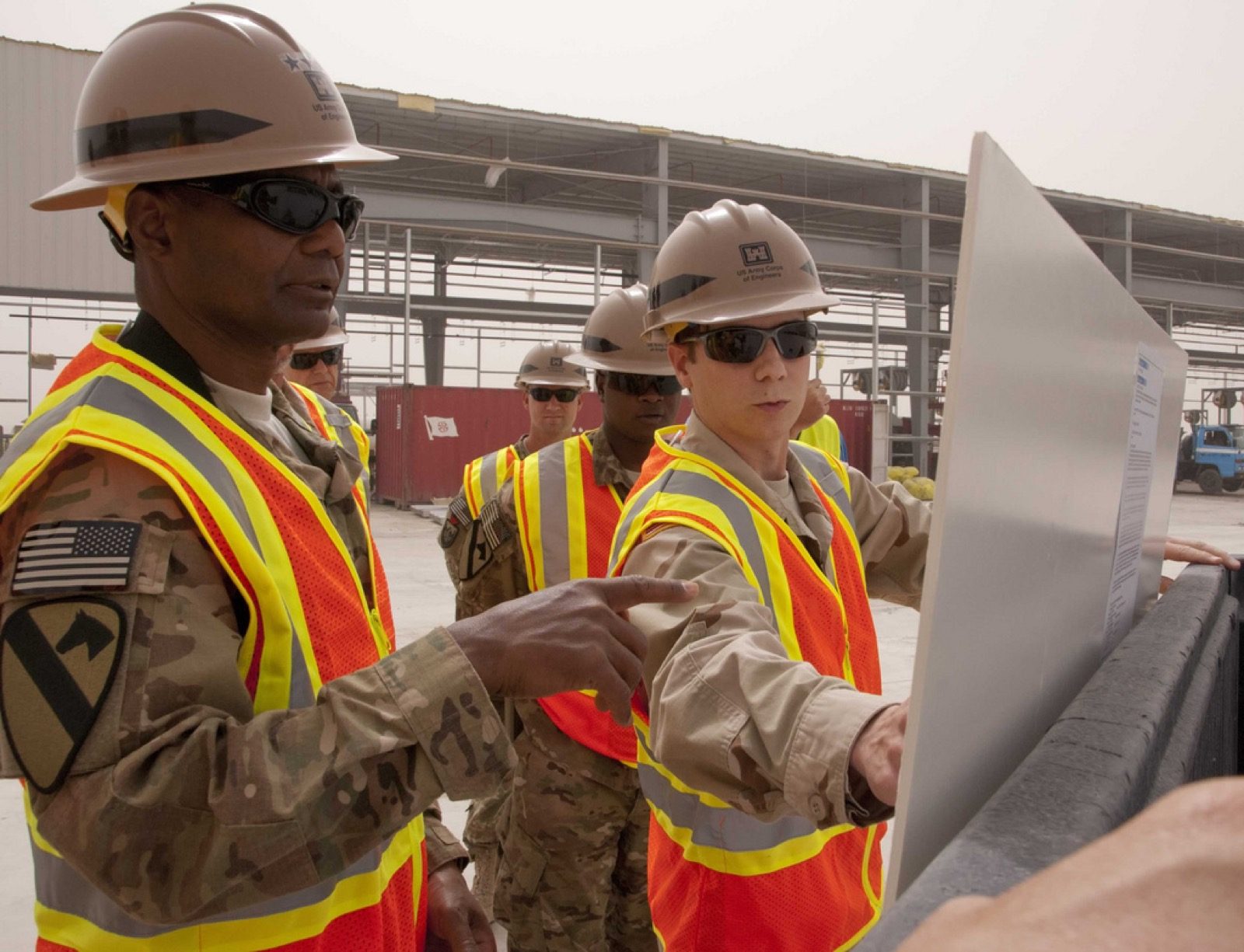 Several people in hard hats and high-visibility vests standing together on a construction site, studying a large drawing sheet propped up in front of them while one points to a detail; the steel frame of the building under construction rises in the background