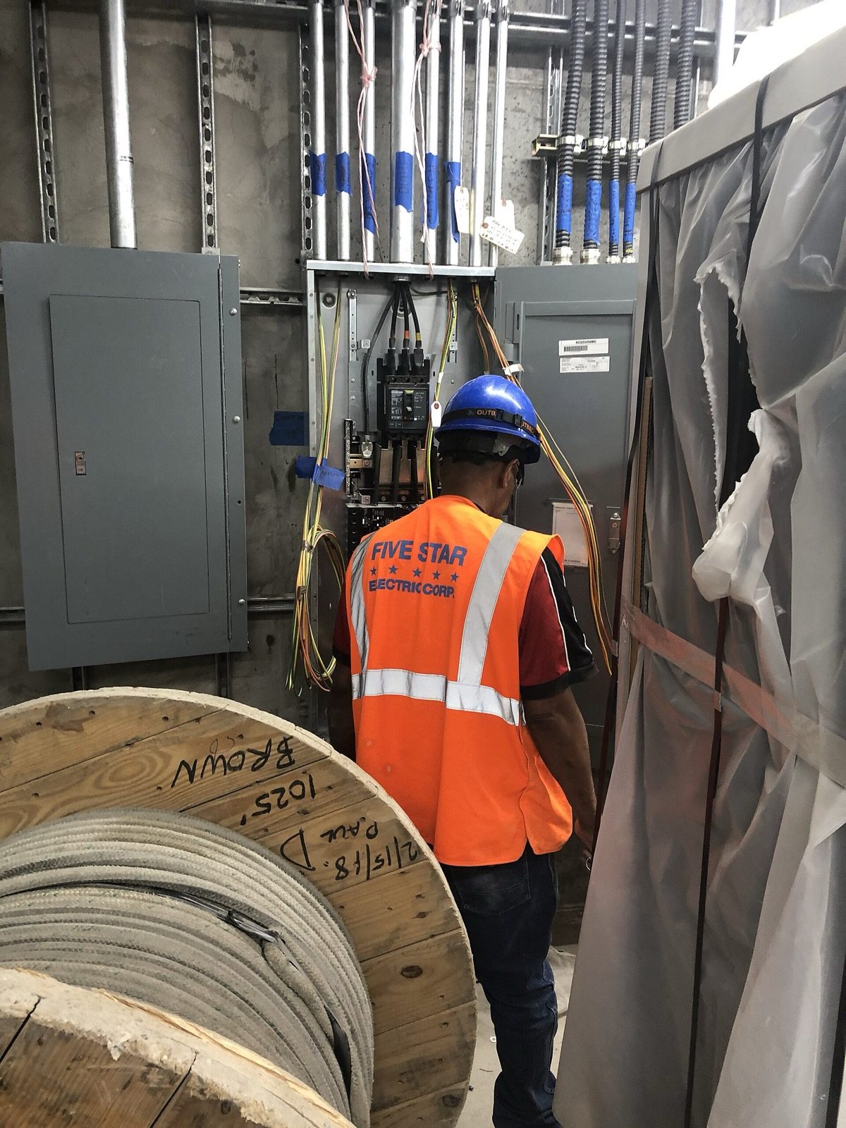 A site photograph of an electrician in high-visibility clothing and a hard hat terminating branch-circuit conductors at an open electrical panel, with colour-coded wires fanned out, conduit risers entering the board from above and a large drum of cable in the foreground