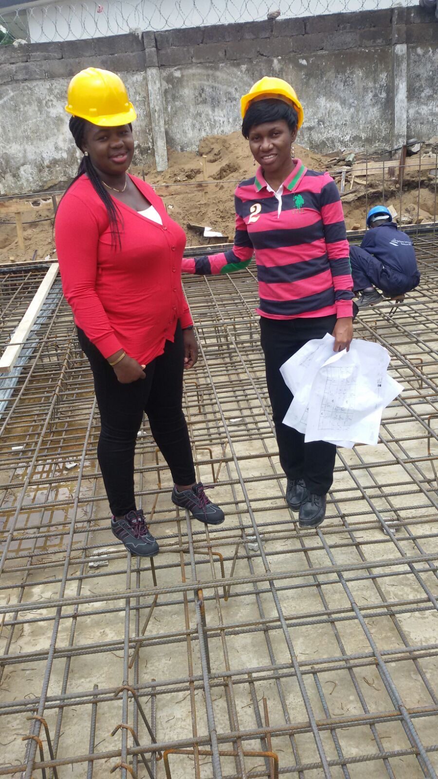 Engineers standing on a tied steel reinforcement mat laid at ground level before concreting, one holding a rolled drawing, showing the grid of bars that a footing schedule specifies as steel inside the foundation