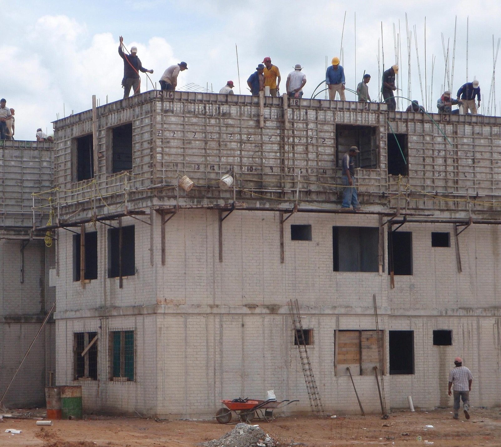 A multi-storey building under construction, its reinforced-concrete frame plainly visible — cast floor slabs, the upright columns and the spanning beams of the upper floor still in formwork, with workers placing reinforcement on the top level; this is the physical skeleton where the architectural, structural and services drawings all meet on one shared grid