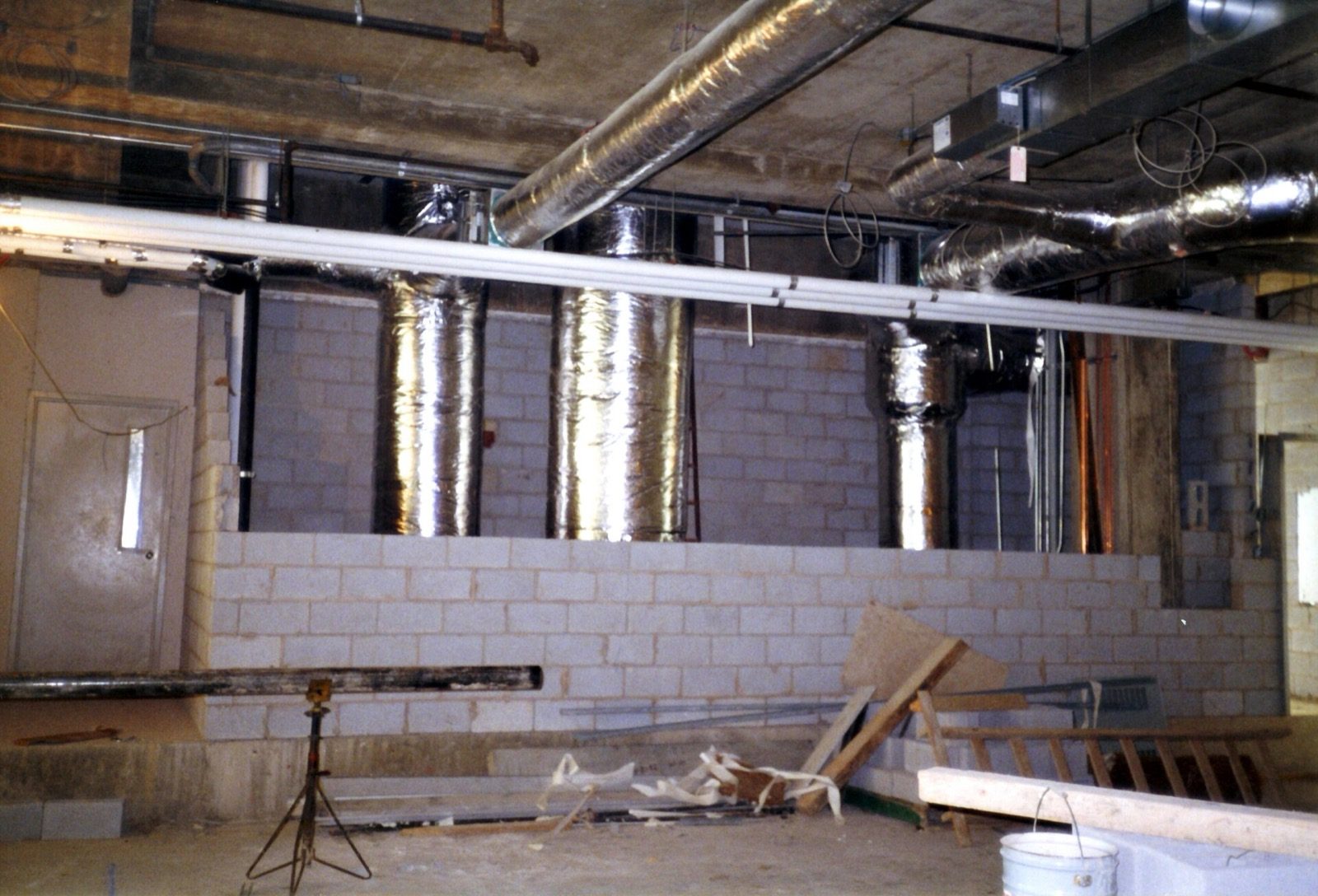 A real building services ceiling void during construction: round insulated metallic supply ducts rising through a concrete shaft alongside bare block walls and service pipes, before the false ceiling is built to hide them