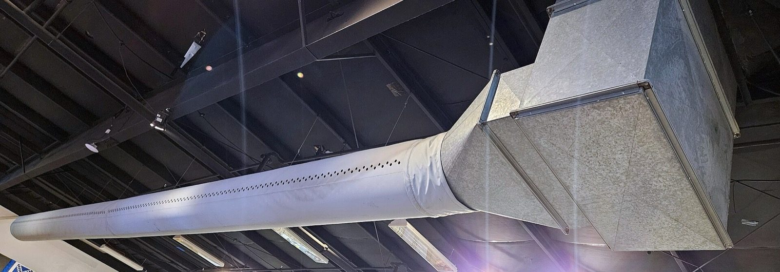 A real installed HVAC duct: a round white insulated flexible-style duct transitioning into a large rectangular galvanised sheet-metal trunk duct, suspended below an exposed structural ceiling — the physical equivalent of the wide double-line channels drawn on the plan