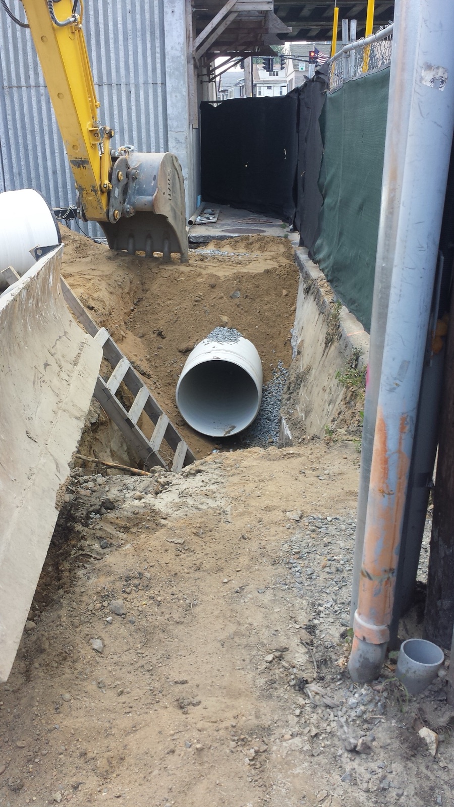 A large-diameter PVC drainage pipe being installed in an open excavated trench, bedded on a gravel haunch near a manhole, with the trench shored at the sides — the kind of heavy gravity drainage line that a plumbing drawing represents as a single heavy line on plan