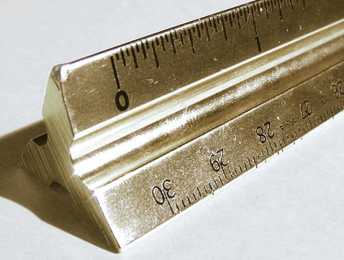 Close-up of a metal triangular architect's scale rule, its three faces each carrying a different graduated scale, with the zero mark and numbered divisions clearly visible