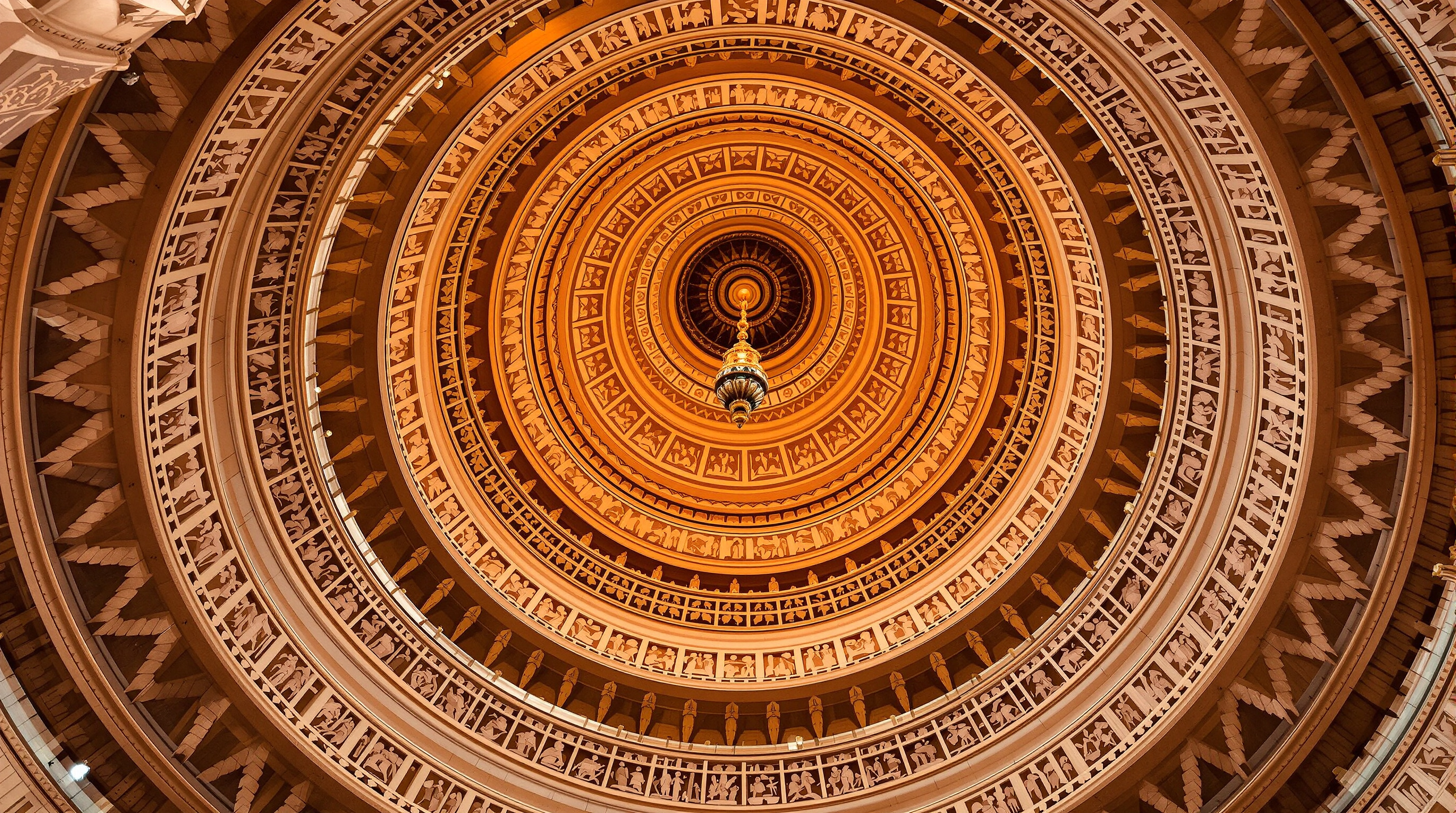 A cluster of Akshardham's corbelled stone domes seen from below, their concentric rings of carved sandstone spiralling inward to a central pendant, every band dense with figures and foliate ornament, warm light raking across the relief