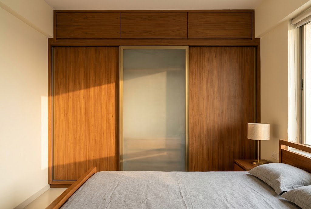 Master bedroom wall in a typical Indian apartment showing a floor-to-ceiling wardrobe of ten foot wide by nine foot tall with three sliding doors — two in Burma teak veneer and one centre door in six millimetre frosted-glass on a brushed brass frame — and an overhead loft storage band eighteen inches high above with three lift-up shutters in matching teak set against a warm cream-painted bedroom wall, a low king-size bed with a clean grey linen bedspread partly visible in the lower foreground, a teak nightstand with a brass table lamp on the right, and warm late-afternoon side light entering from a window beyond on the right casting a soft sunlit band on the wardrobe doors
