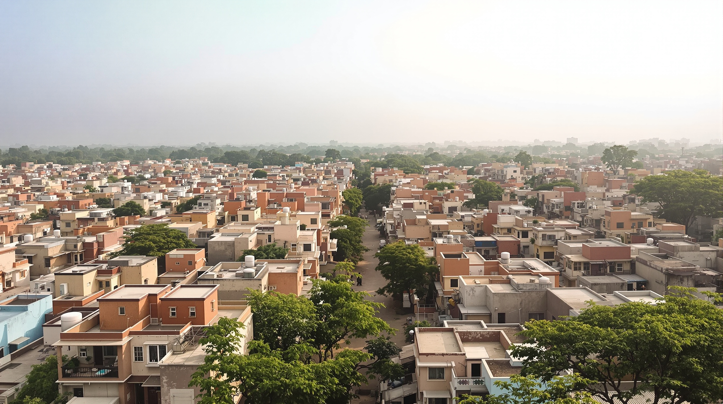 An aerial view of a densely built Aranya sector in Indore: a fabric of flat-roofed two- and three-storey houses in earthen and pastel colours, punctuated by small courtyards and rooftop terraces with water tanks, threaded by a hierarchy of narrow lanes opening onto a wider tree-lined spine