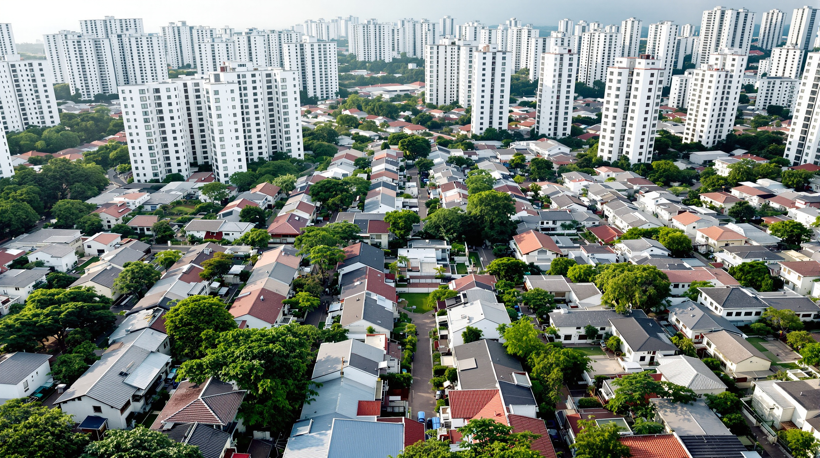 An aerial or elevated view of the Belapur housing district today, the original low-rise white cottages surrounded and dwarfed by newer mid-rise and high-rise apartment towers, with the community courtyards still legible between the older roofs