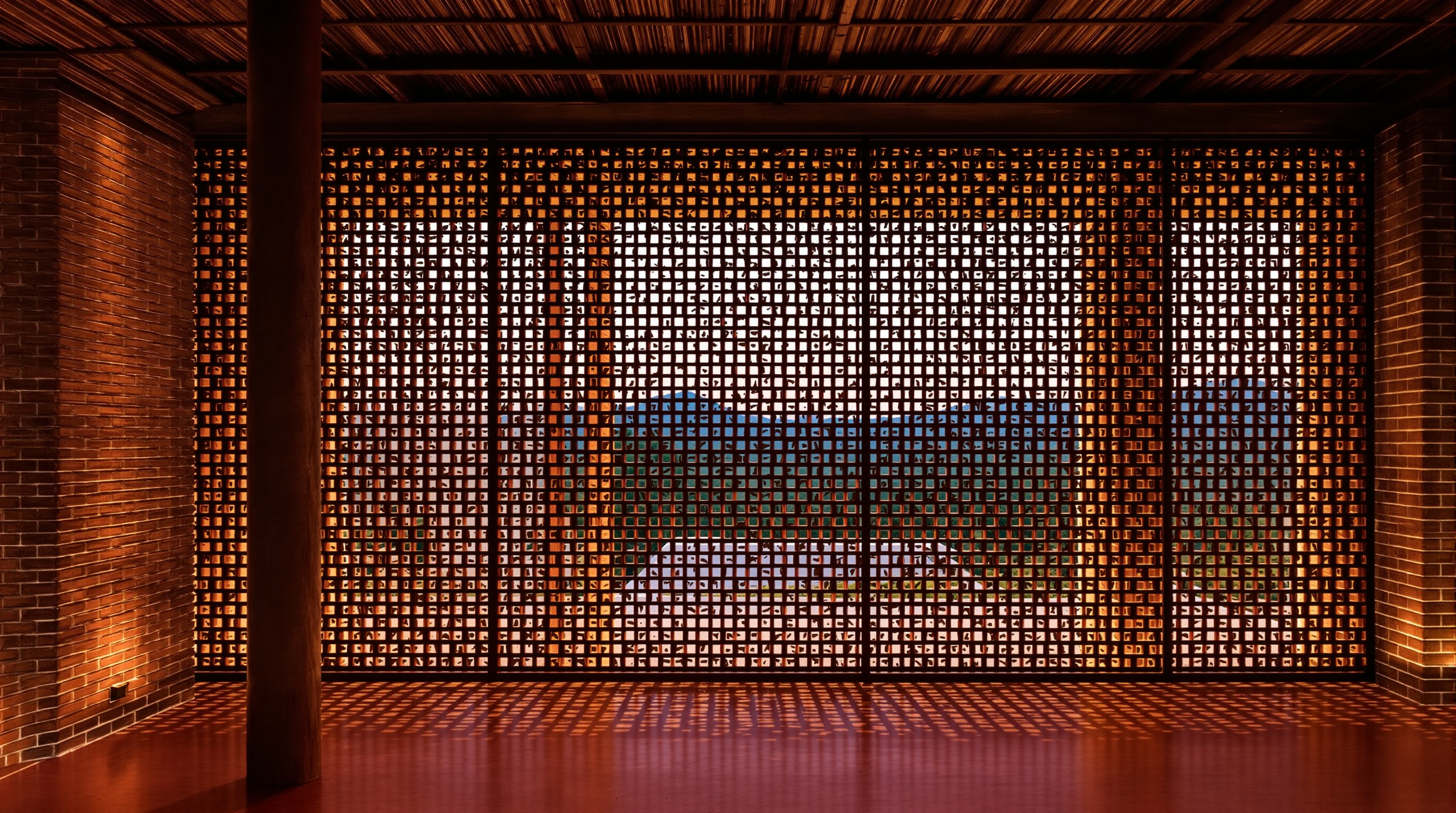 A pierced brick jali wall of the Brick House seen from inside at dusk, the omitted bricks casting a lattice of warm light and shadow across a red-oxide floor, a bamboo-slat ceiling and a timber column visible in the foreground