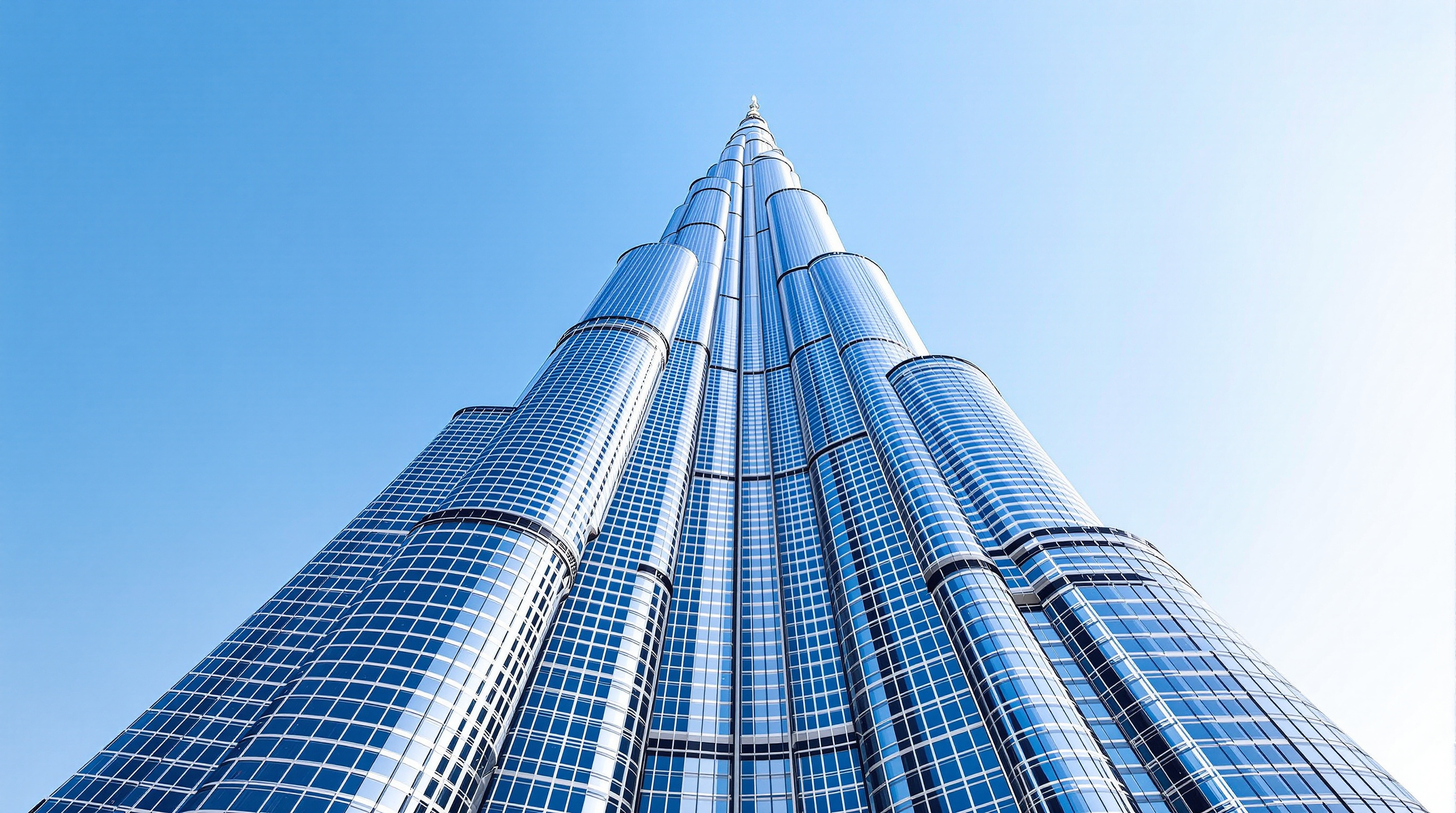 Looking straight up the Burj Khalifa's tapering shaft from its base, the three wings stepping back in a spiralling sequence of silver setbacks against a bright sky, the central spire vanishing to a point