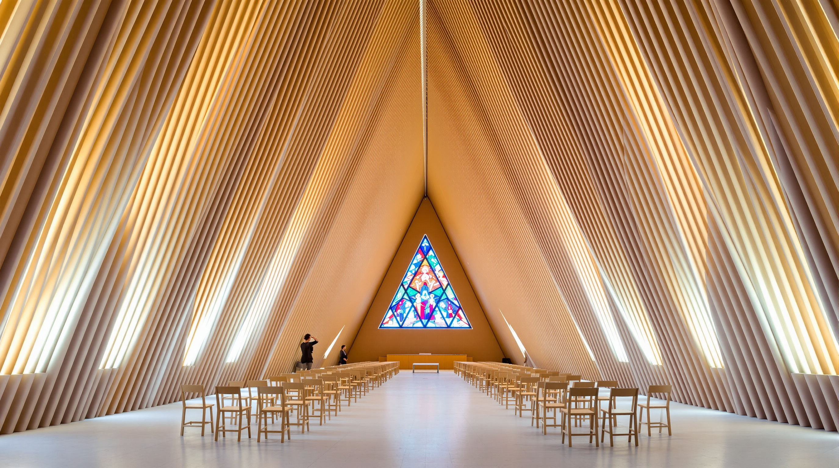 Interior of the Cardboard Cathedral looking toward the altar: the nave rises in a steep triangular vault lined with rows of pale cardboard tubes, daylight glowing through the gaps between them, simple wooden chairs arranged on a bare floor, the triangular stained-glass window casting coloured light at the far end