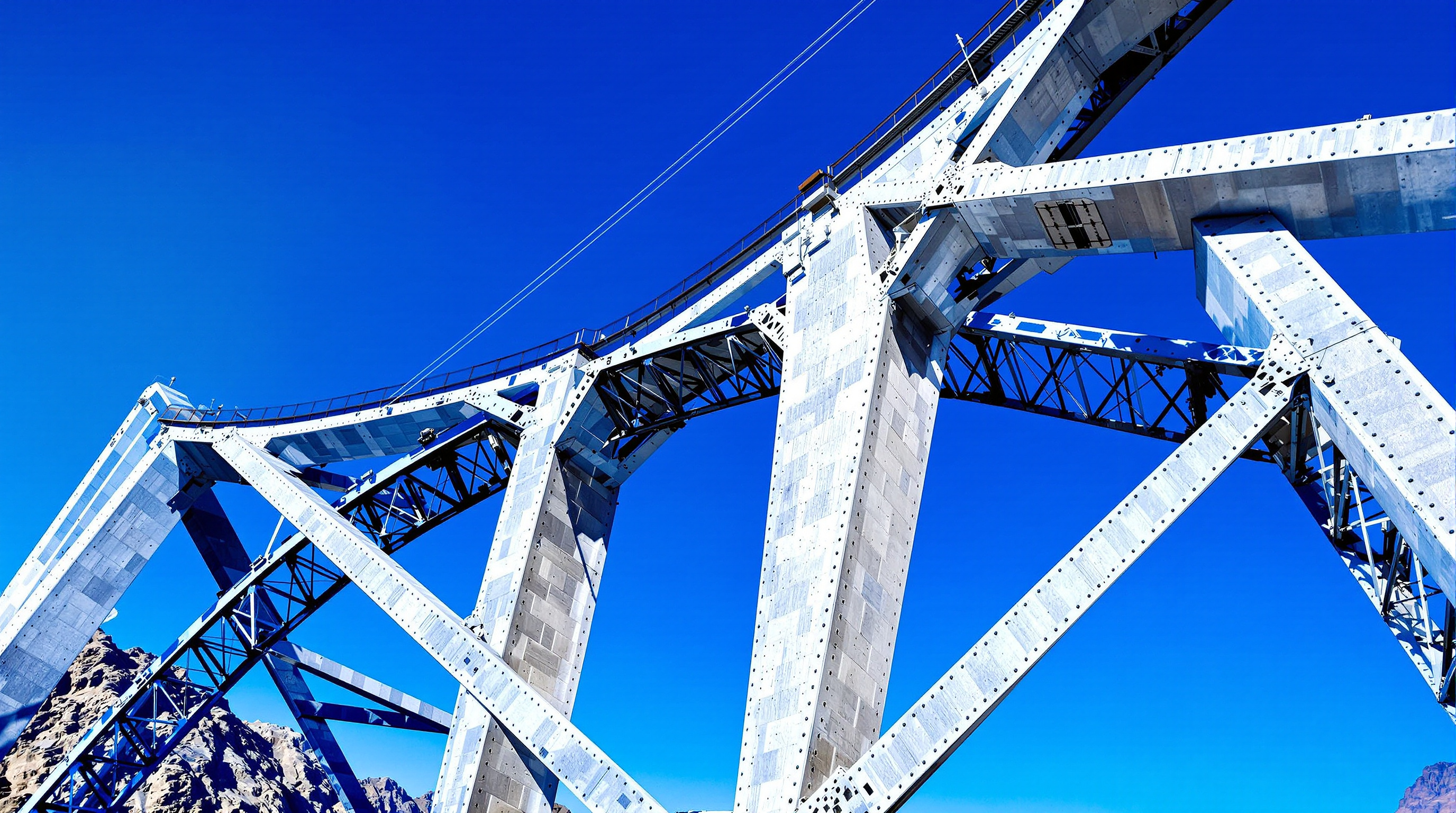 Close view of the twin concrete-filled steel ribs of the Chenab bridge arch meeting at the crown high above the gorge, the riveted silver-grey box sections and cross-bracing catching hard mountain sunlight against a deep blue sky