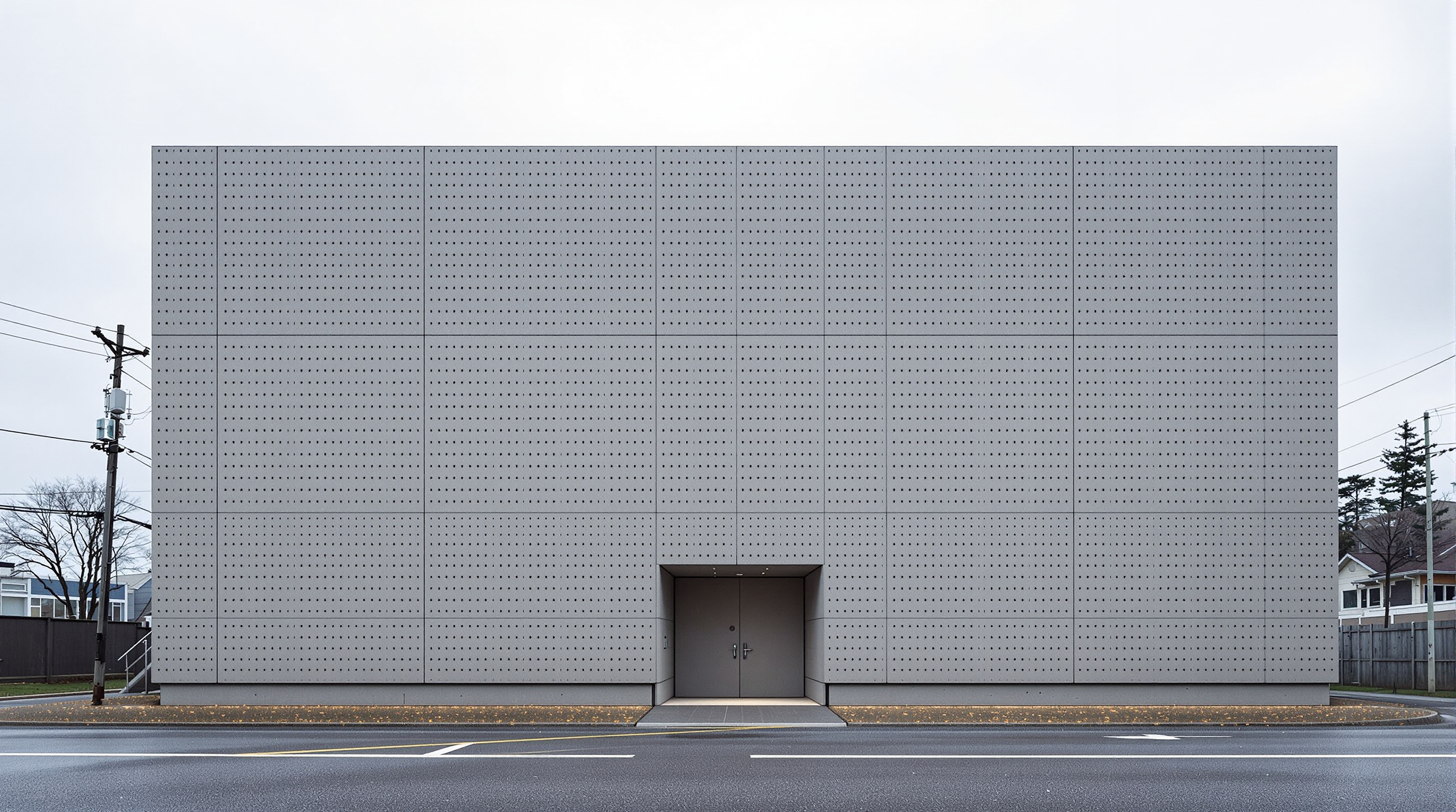 Exterior of the Church of the Light: a stark, windowless grey concrete box in a quiet suburban lot in Ibaraki, its smooth formwork surface marked by a regular grid of small tie-holes, a single low entrance, under an overcast Osaka sky
