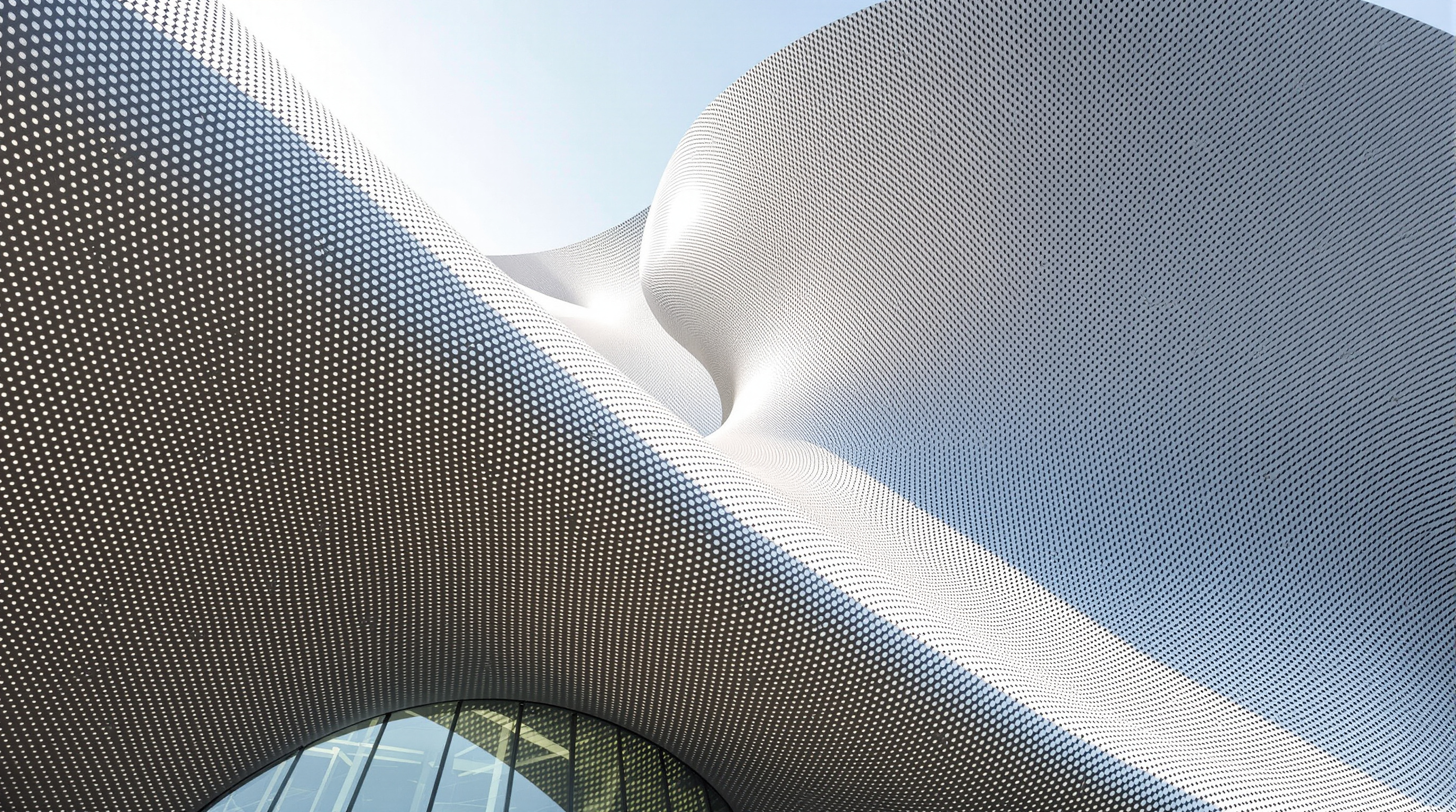 Detail of the Dalian conference centre's outer envelope: thousands of perforated silver-grey aluminium slats curving across a bulging steel form, daylight filtering through the small round holes to speckle the surface, the metal skin folding around a swelling roof volume