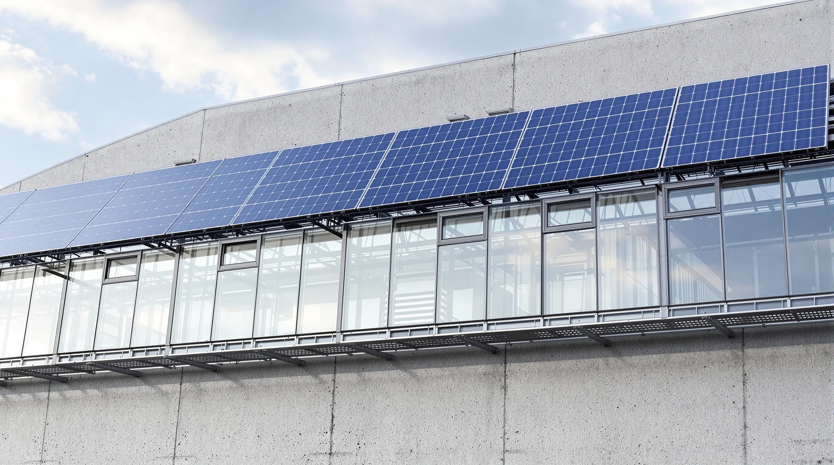 The dark solar skin on the south facade of the Energy Bunker seen close up: rows of glazed solar thermal collectors and blue photovoltaic panels mounted on a steel frame standing proud of the raw grey concrete wall behind, with the sky reflected in the glass