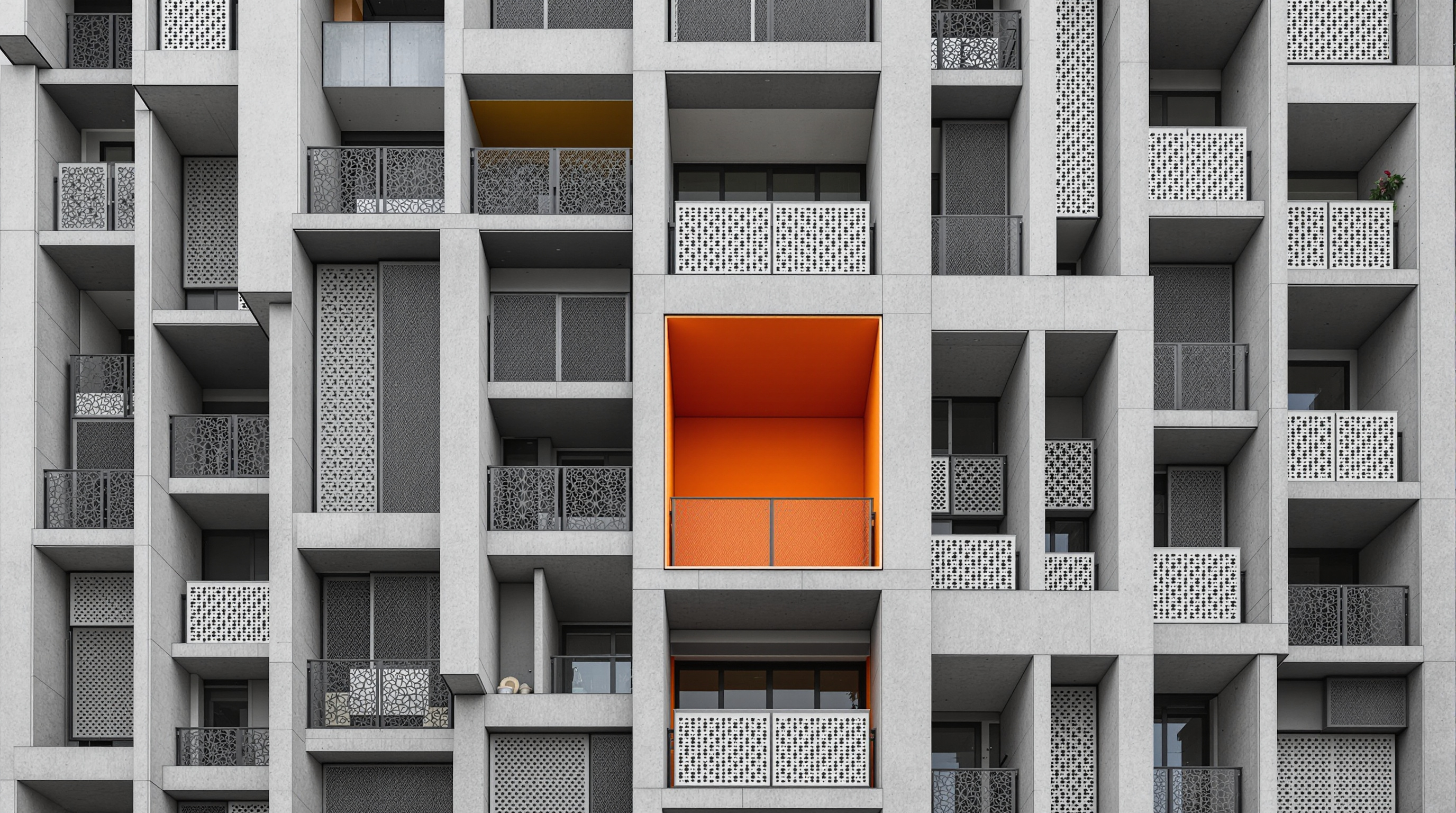 Detail of the Future Towers facade in Pune: deep concrete-framed recessed balconies of varying sizes, some double-height, screened by ornamented perforated-metal shutters, with a bright orange scooped opening puncturing the grey mass