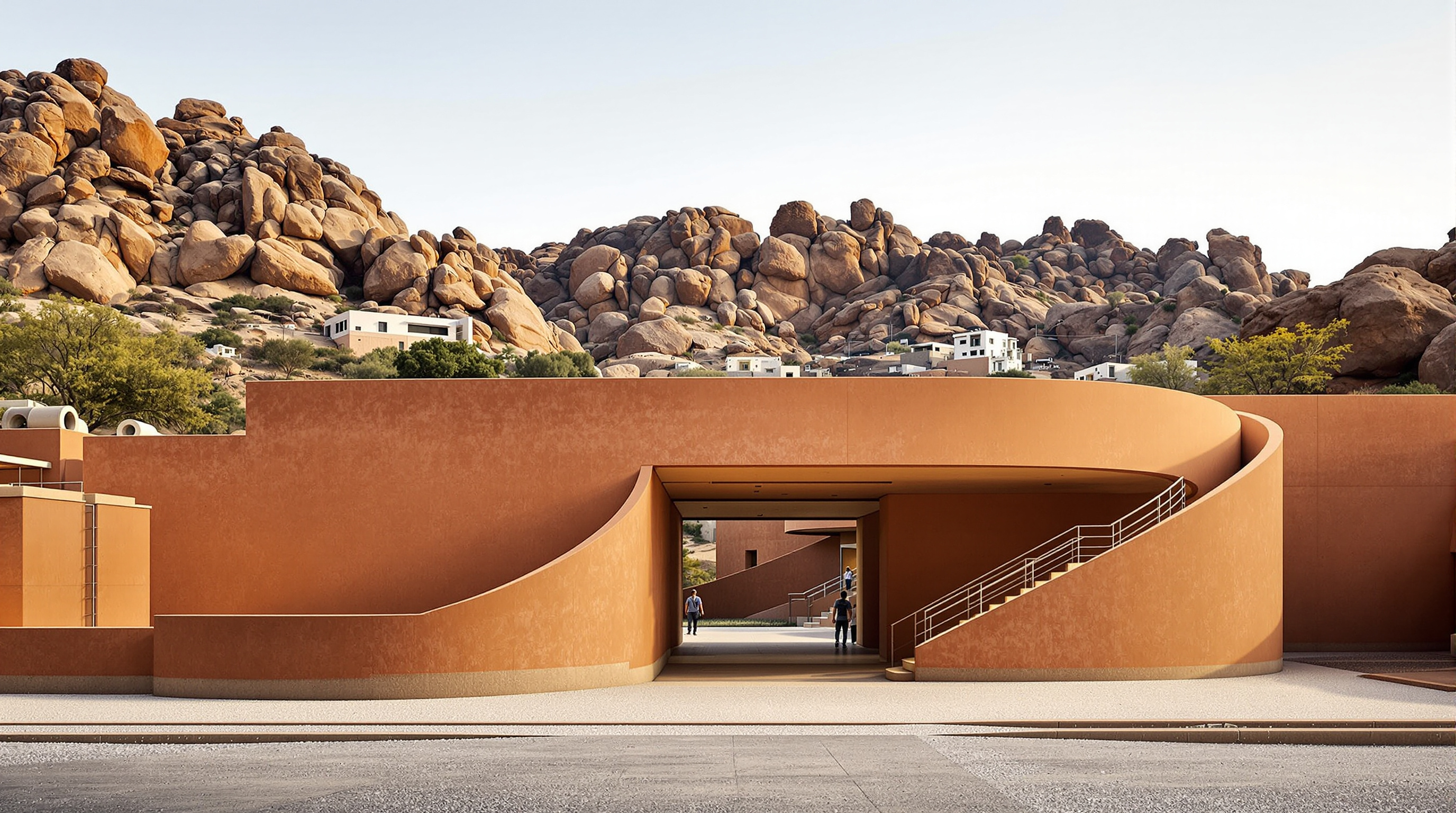 The main entrance approach to Hampi Art Labs, a low curving concrete wall in warm terracotta tones leading visitors along a stabilised-pebble path toward a shaded threshold, boulder-strewn Hampi hills rising behind