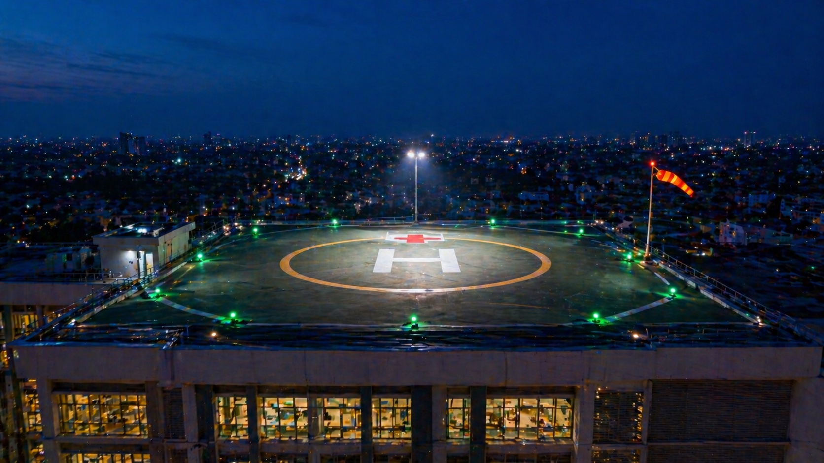 Hospital rooftop helipad at twilight — perimeter green lights, illuminated wind sock, white floodlit H letter, IFR-capable