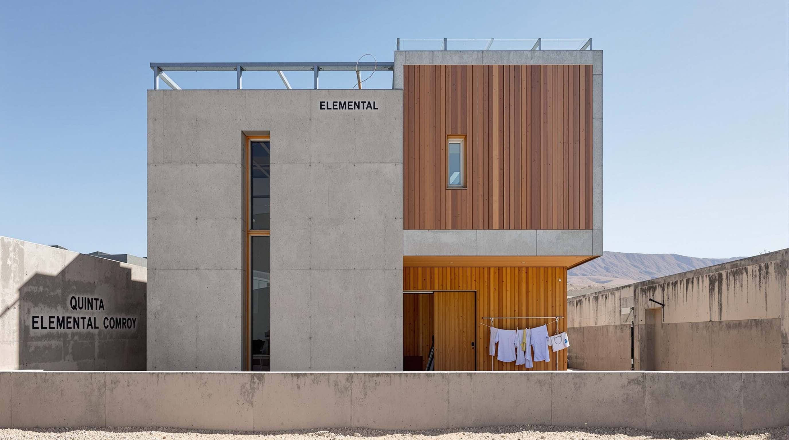 A single Quinta Monroy dwelling photographed close up, its original grey concrete frame clearly visible on one side and a resident-built extension of warm timber cladding and a new window filling the adjacent bay, laundry hanging in the courtyard, the two halves of one house sitting side by side