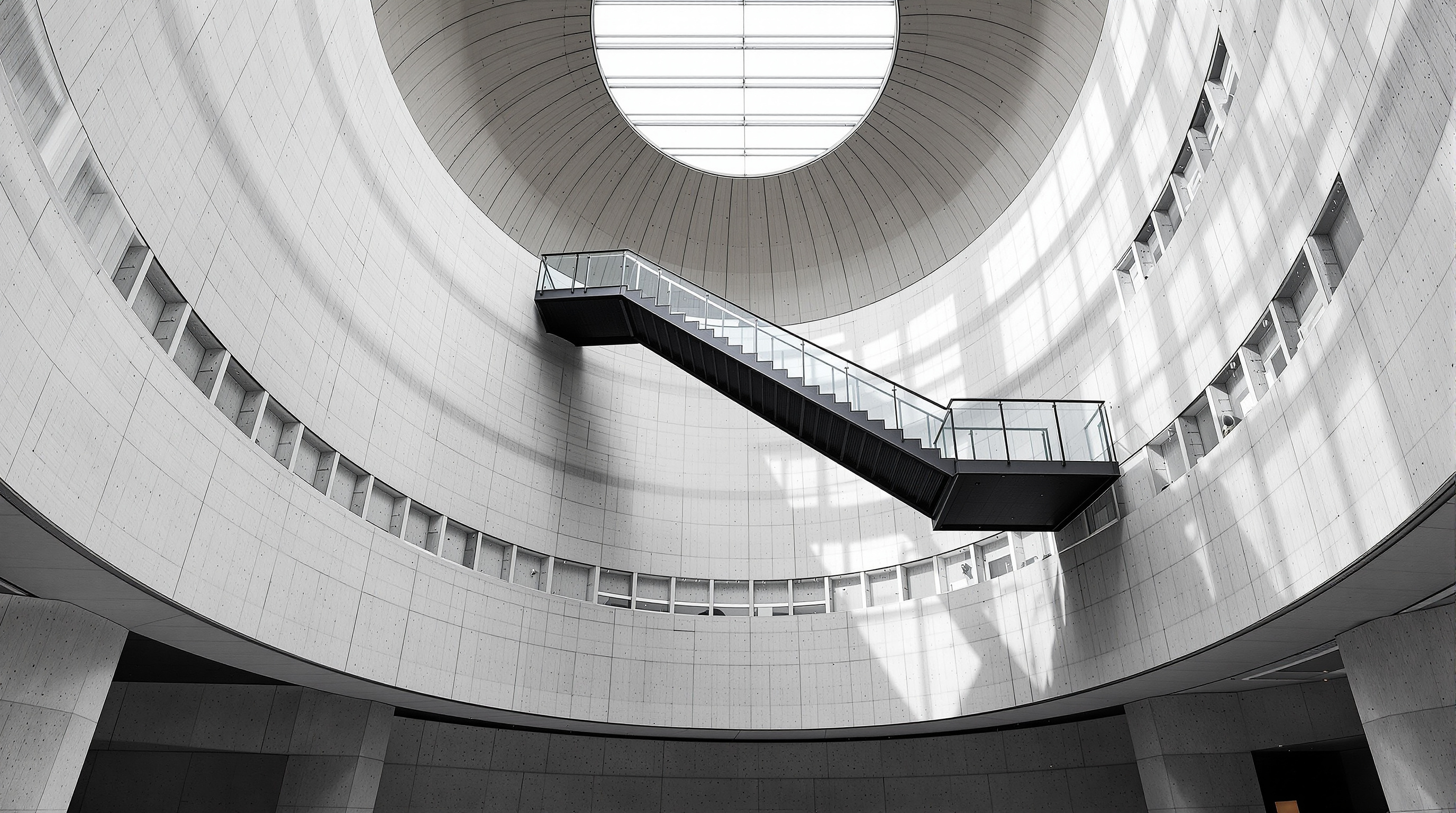 The soaring interior of MAXXI: tall curving grey concrete gallery walls sweeping overhead, a dramatic suspended black steel staircase cutting diagonally through the central void, and a ceiling of linear skylights washing the space with soft even daylight