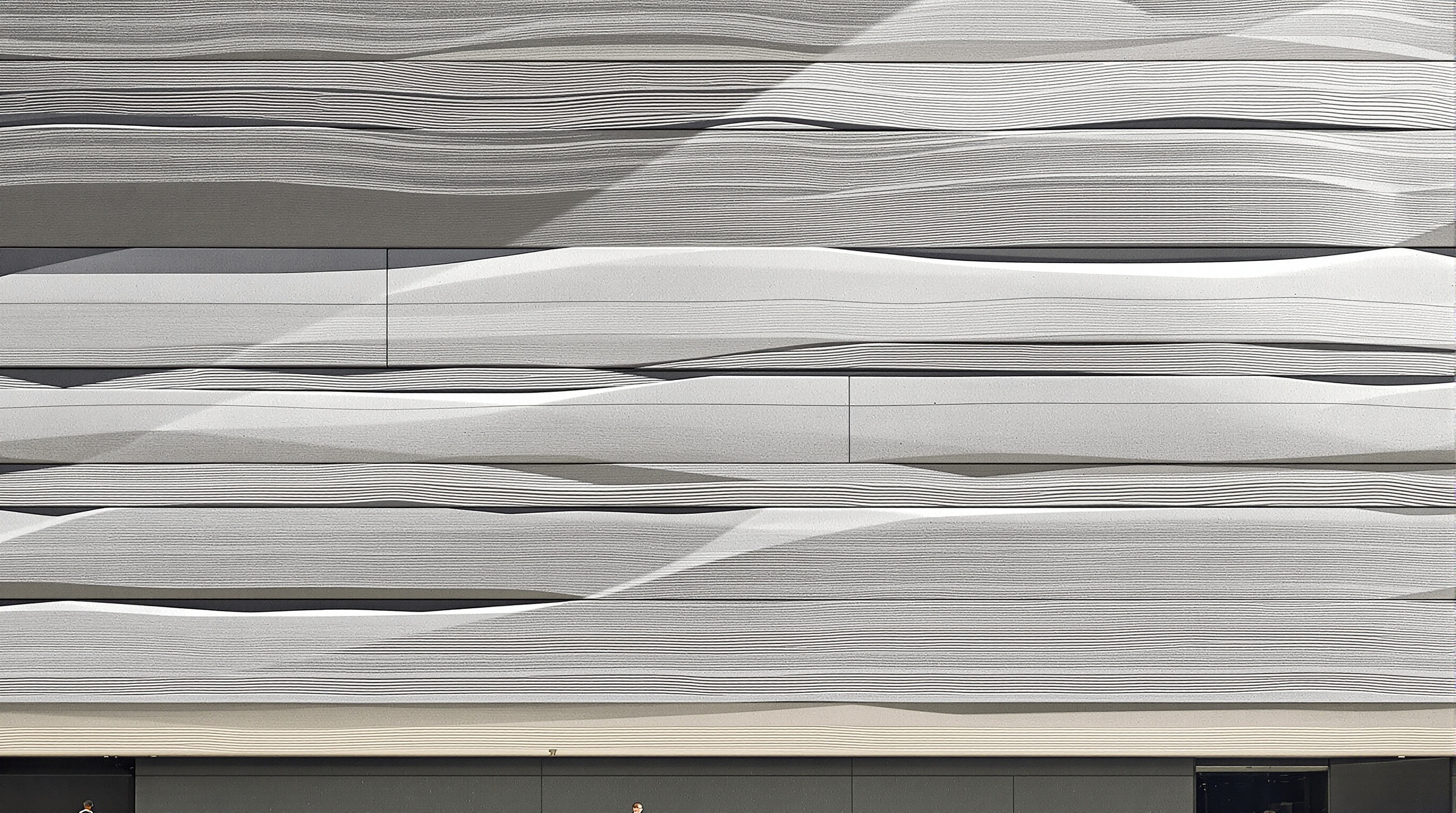 Close-up of the Perot Museum's precast concrete facade: massive grey panels with deep horizontal ribs and striations resembling layers of sedimentary rock, the shadow lines sharp in raking sunlight, a small human figure at the base for scale