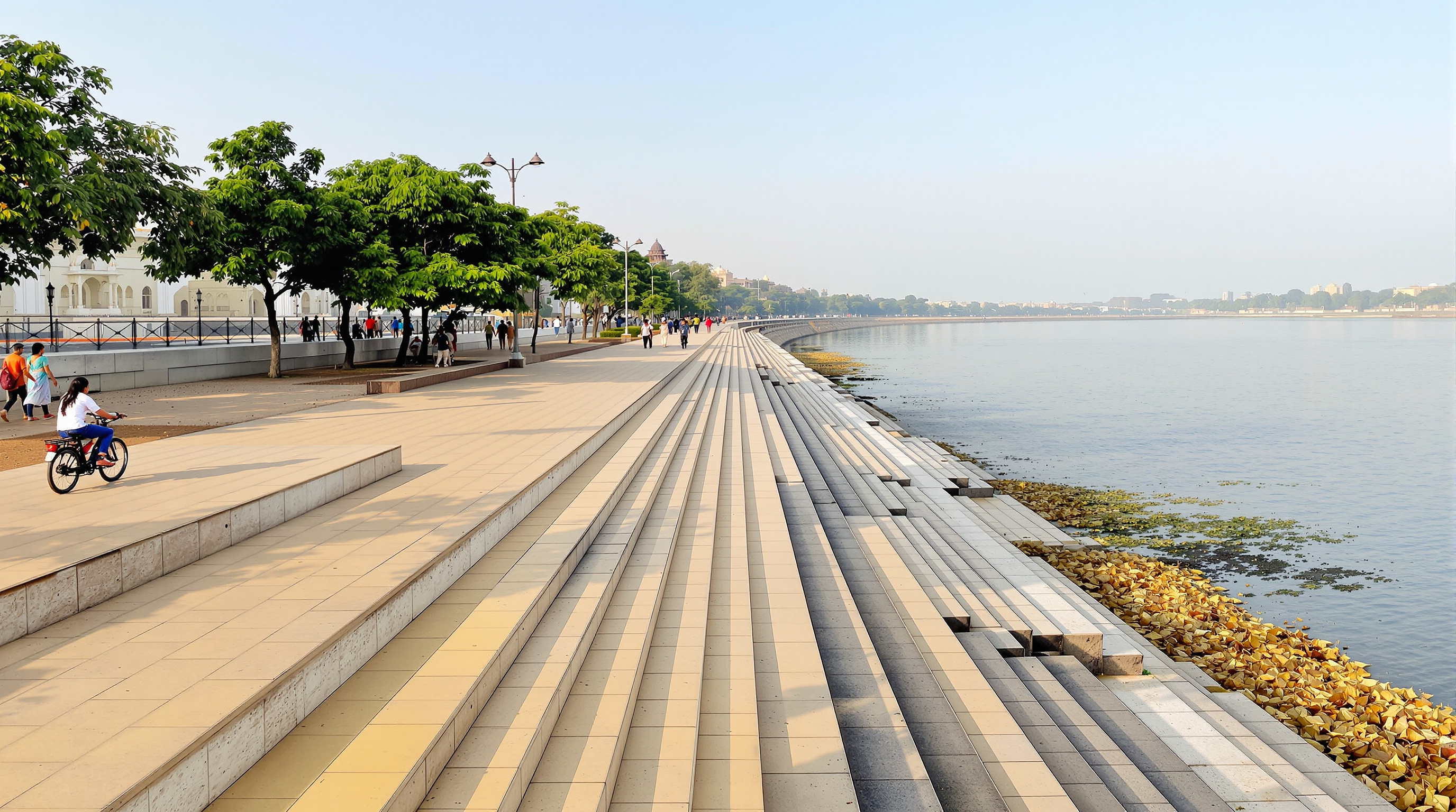 The continuous lower promenade of the Sabarmati Riverfront: broad tan stone paving beside the still water, a low parapet at the edge, people walking and cycling, wide steps (ghats) descending to the water, the flood-retaining wall reading as a clean architectural line