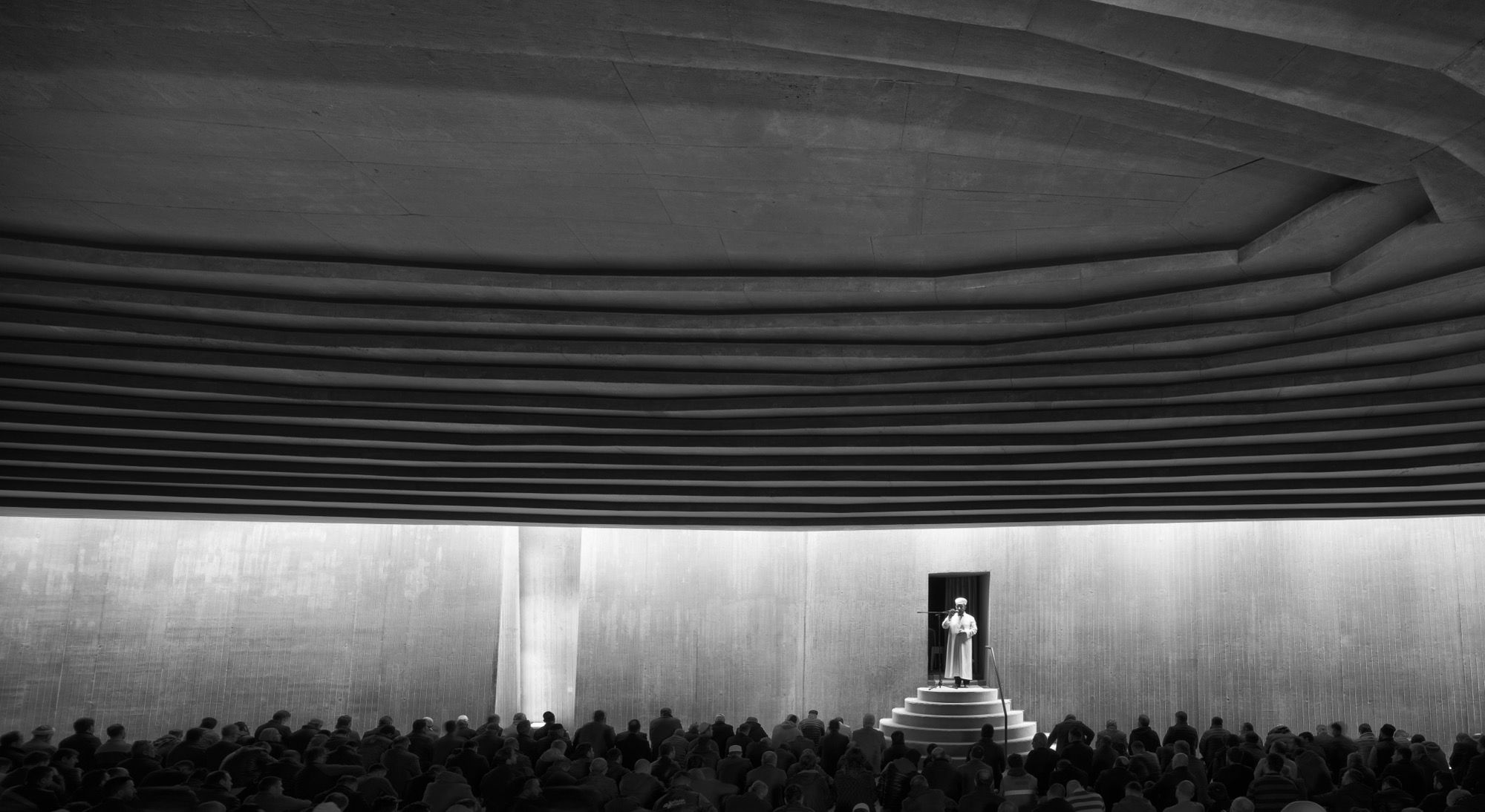 Friday prayers: the imam delivers the sermon from the stepped minbar as the congregation sits across the descending floor, the ribbed concrete ceiling and the luminous qibla wall framing the gathering