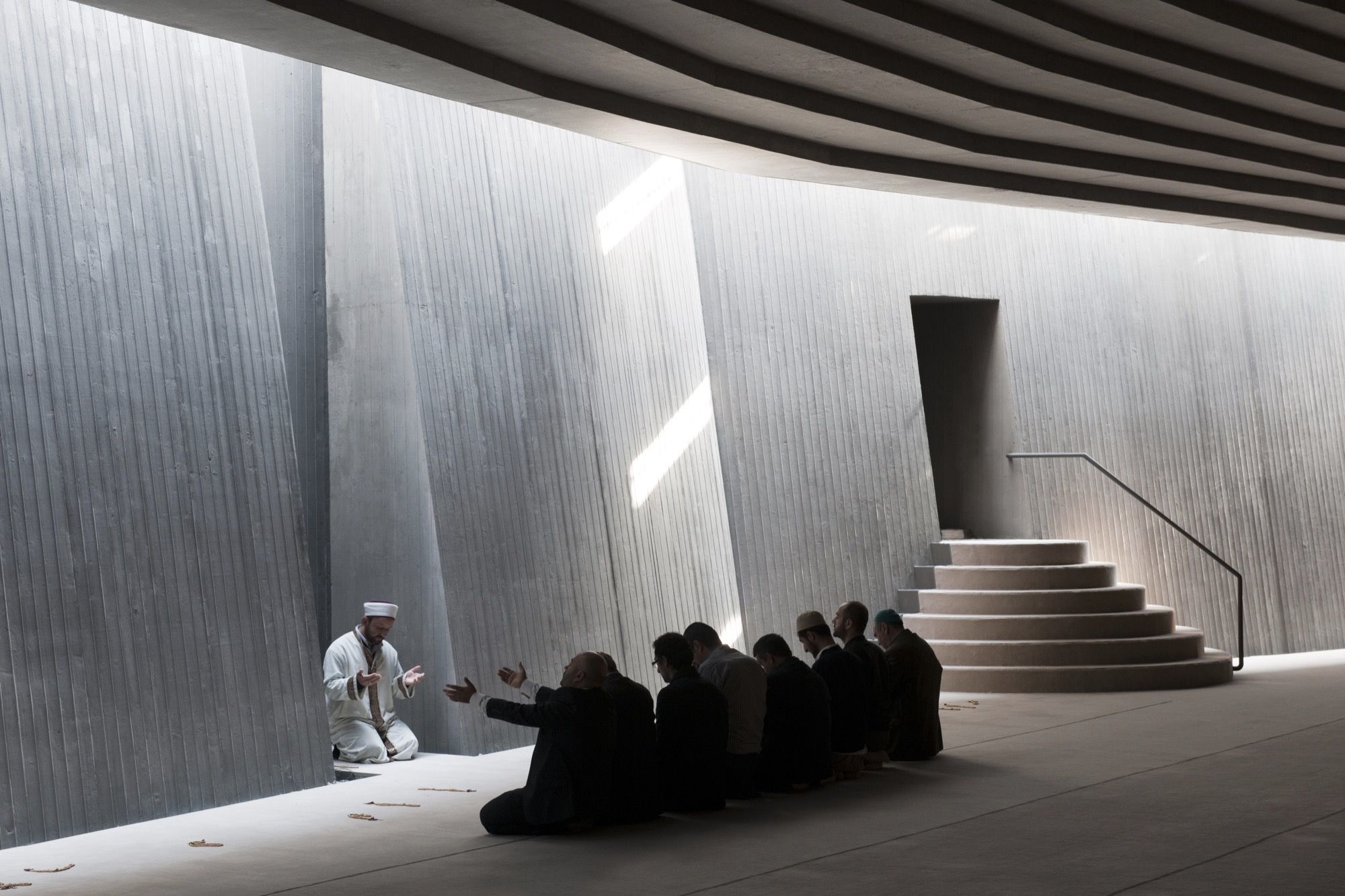 A smaller gathering: the imam kneels and speaks, hands open, to a handful of worshippers seated along the board-marked wall, daylight falling in soft blades and the circular minbar and dark niche to the right