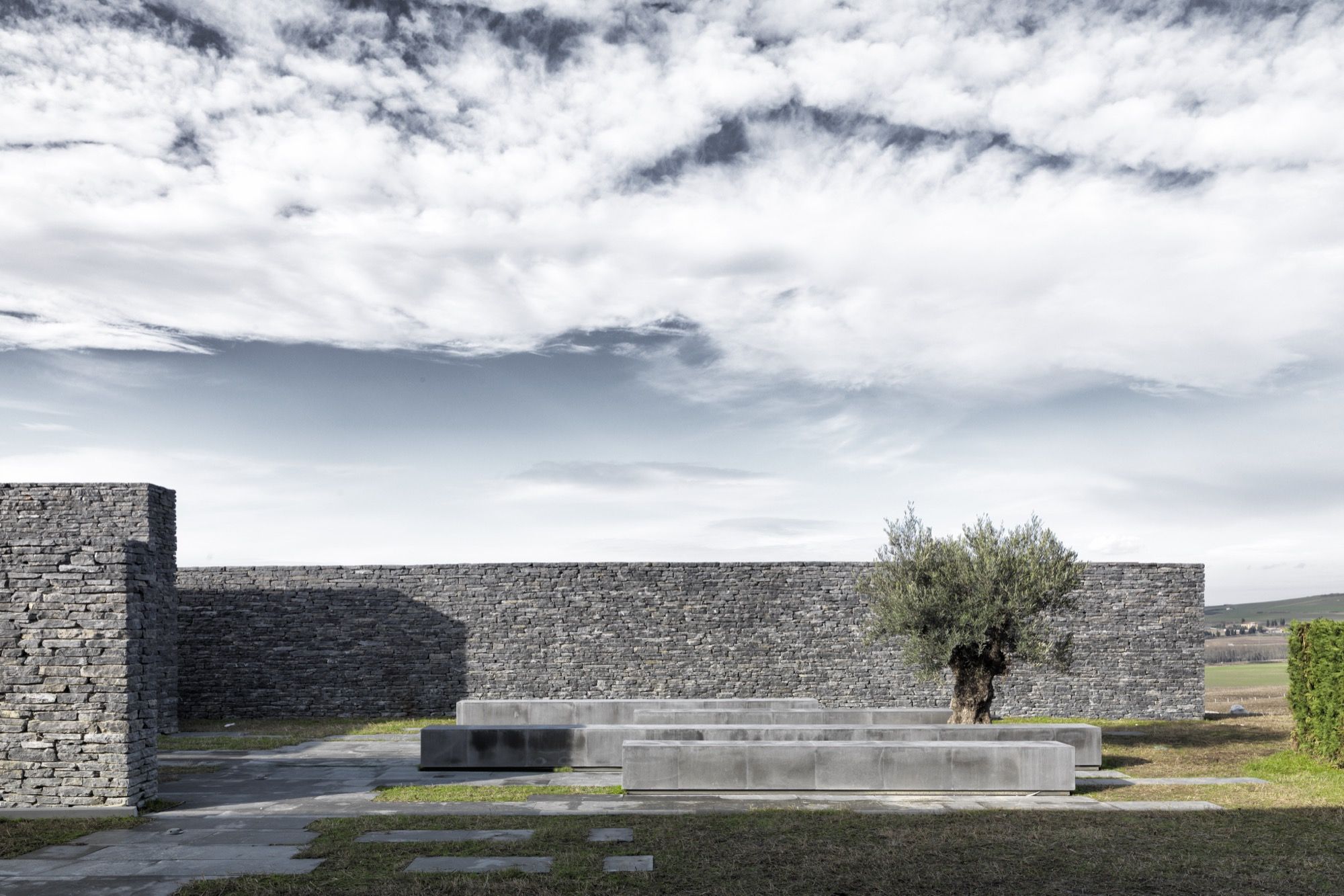 An enclosed court of split-stone walls, a single cast-concrete bench and an old olive tree, open to the sky and the surrounding fields — the mosque's quiet outdoor room