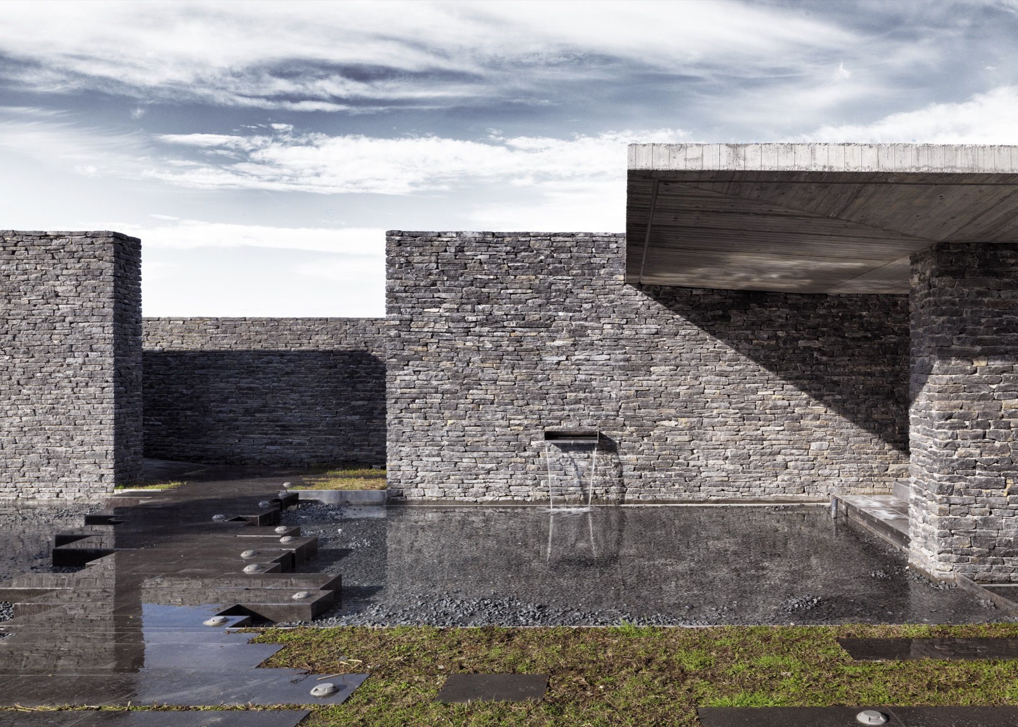 Water and stone in the forecourt: a thin sheet of water slips from a spout in the split-stone wall into a dark reflecting pool set with stepping stones, the cantilevered roof throwing shade at the right