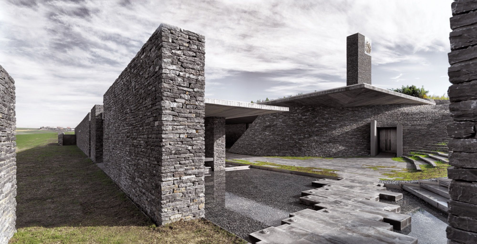 Split-stone walls fold across the forecourt toward the entrance, a shallow water channel and stepping pavers threading between them beneath the cantilevered roof and the calligraphic-topped minaret