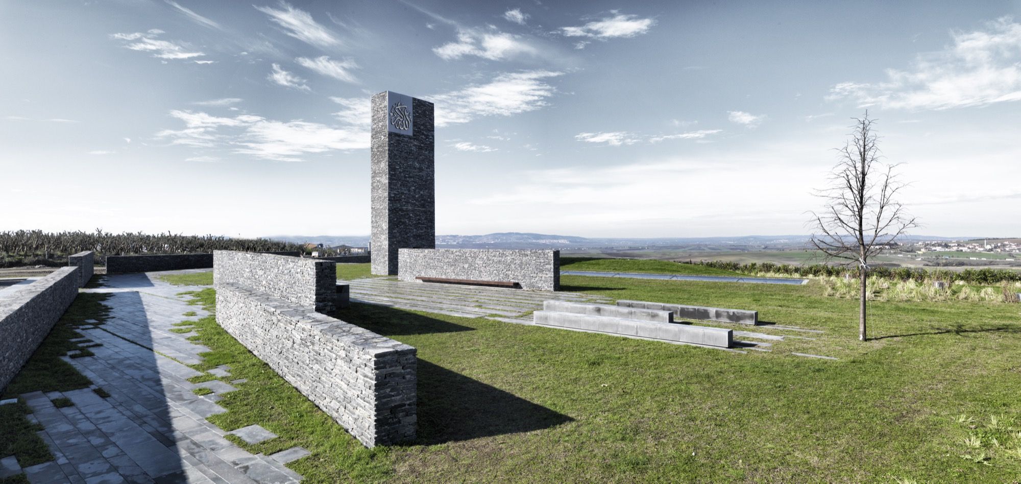 On the planted roof terrace the plain split-stone minaret — its only marking a pierced calligraphic panel near the top — stands over low stone walls, cast-concrete benches and a young tree, the Thracian plain running to the horizon