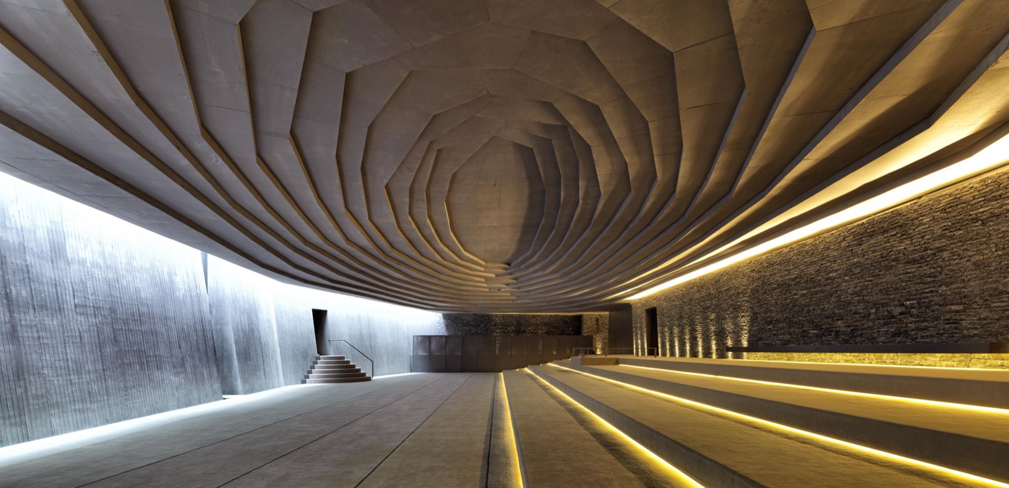 Inside the prayer hall: the board-marked concrete ceiling fans inward in stepped rings like the roof of a geode, grazing daylight and a hidden light-line washing down the split-stone qibla wall on the right while the plain floor steps down toward Mecca