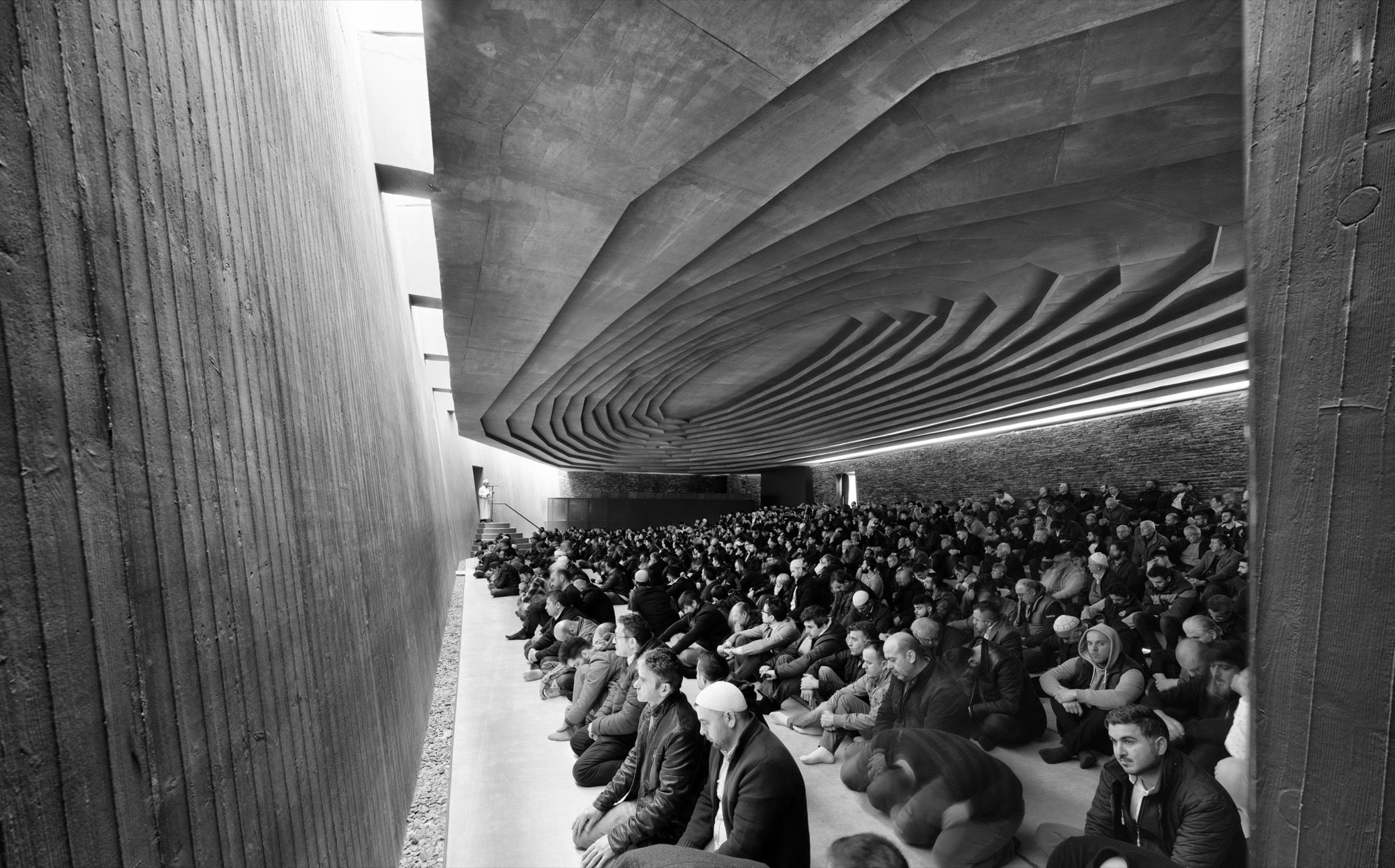 A full congregation at prayer in the low, cave-like hall, seated on the stepped stone floor beneath the banded concrete ceiling, the board-marked side wall to the left and the imam small at the distant minbar