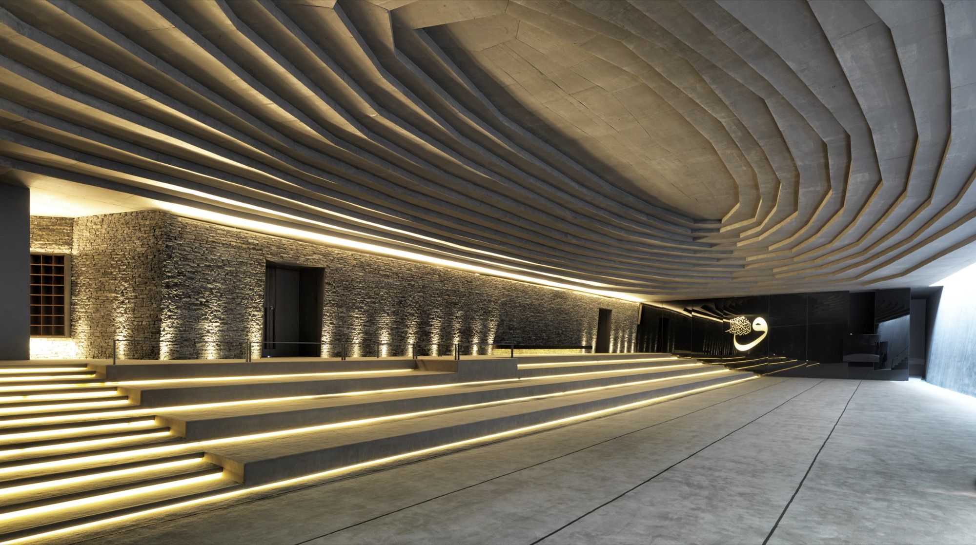The hall seen from the rear: concealed light-lines trace every step of the concrete ceiling and floor, the rough qibla wall glowing at the left, while a single illuminated line of Arabic calligraphy floats on the dark polished wall to the right