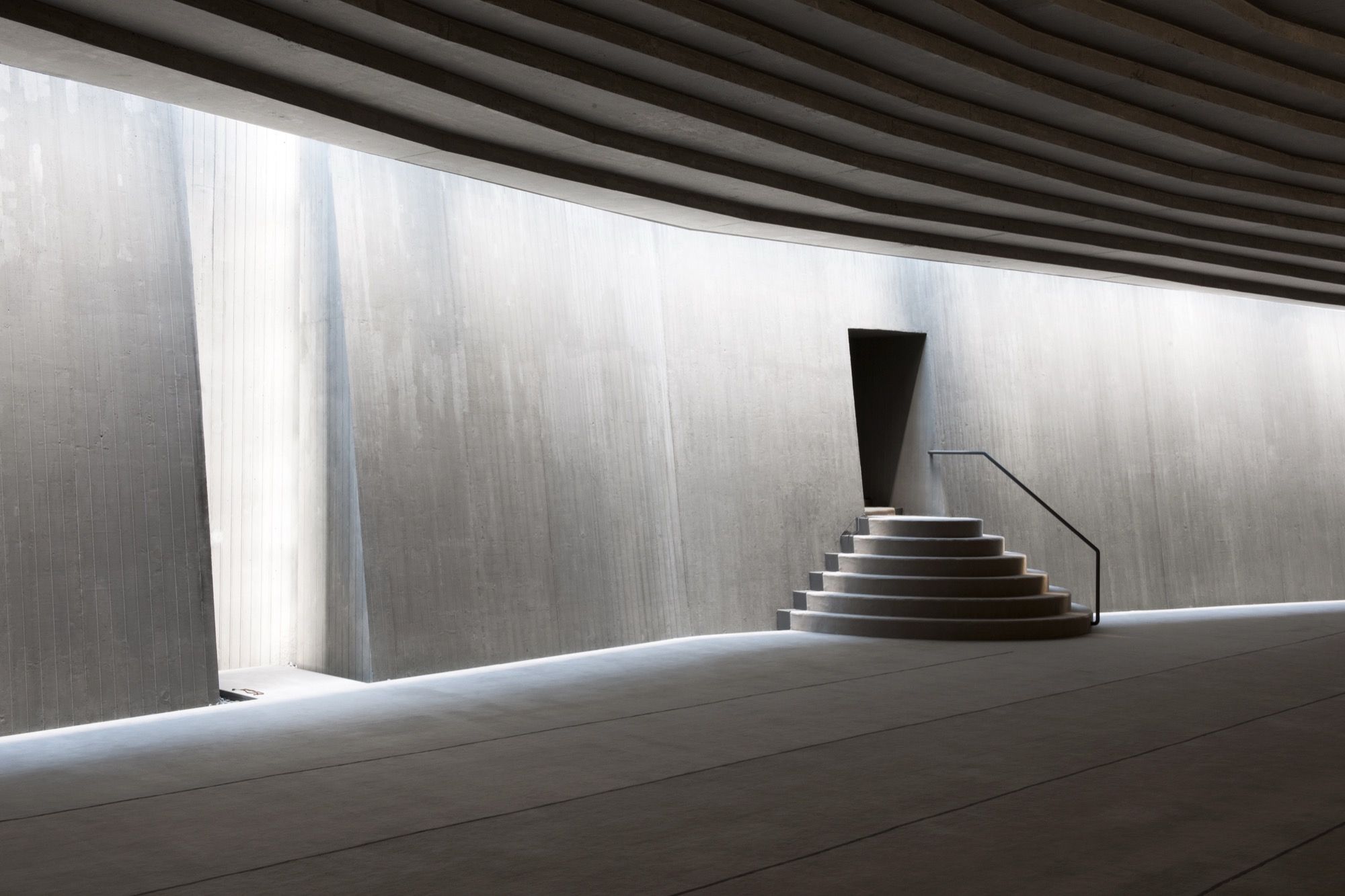 The minbar — a simple circle of concrete steps with a slim steel rail — set against the curving board-marked wall beneath a narrow tapering niche, lit only by the soft daylight sliding down the concrete