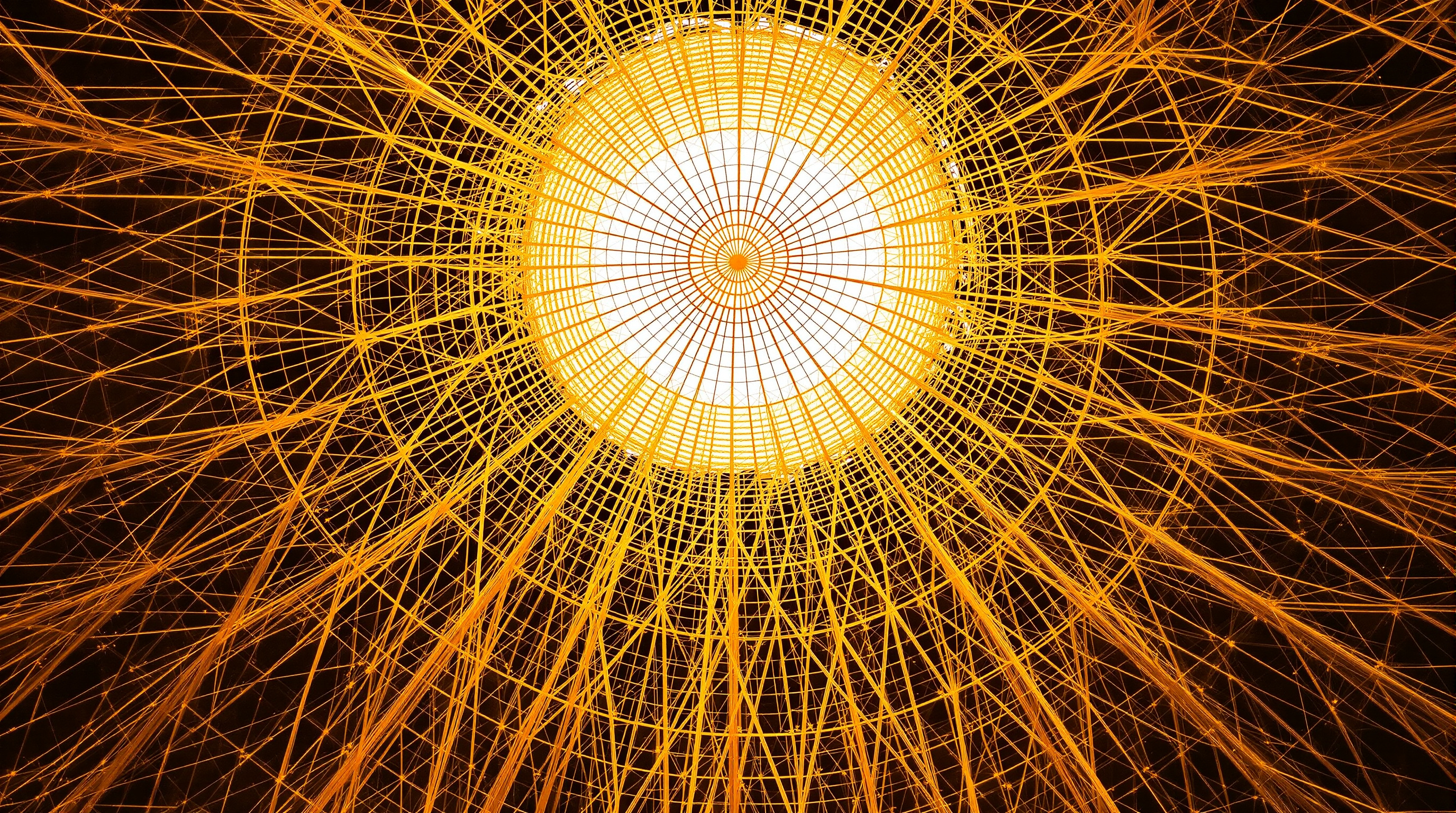 The completed Silk Pavilion viewed from below, looking up into the hemispherical dome: an intricate web of amber and white silk, the geometric CNC-laid lattice radiating from the crown, softened everywhere by the cloudy non-woven silk the worms have spun between the structural threads, backlit so the whole surface glows
