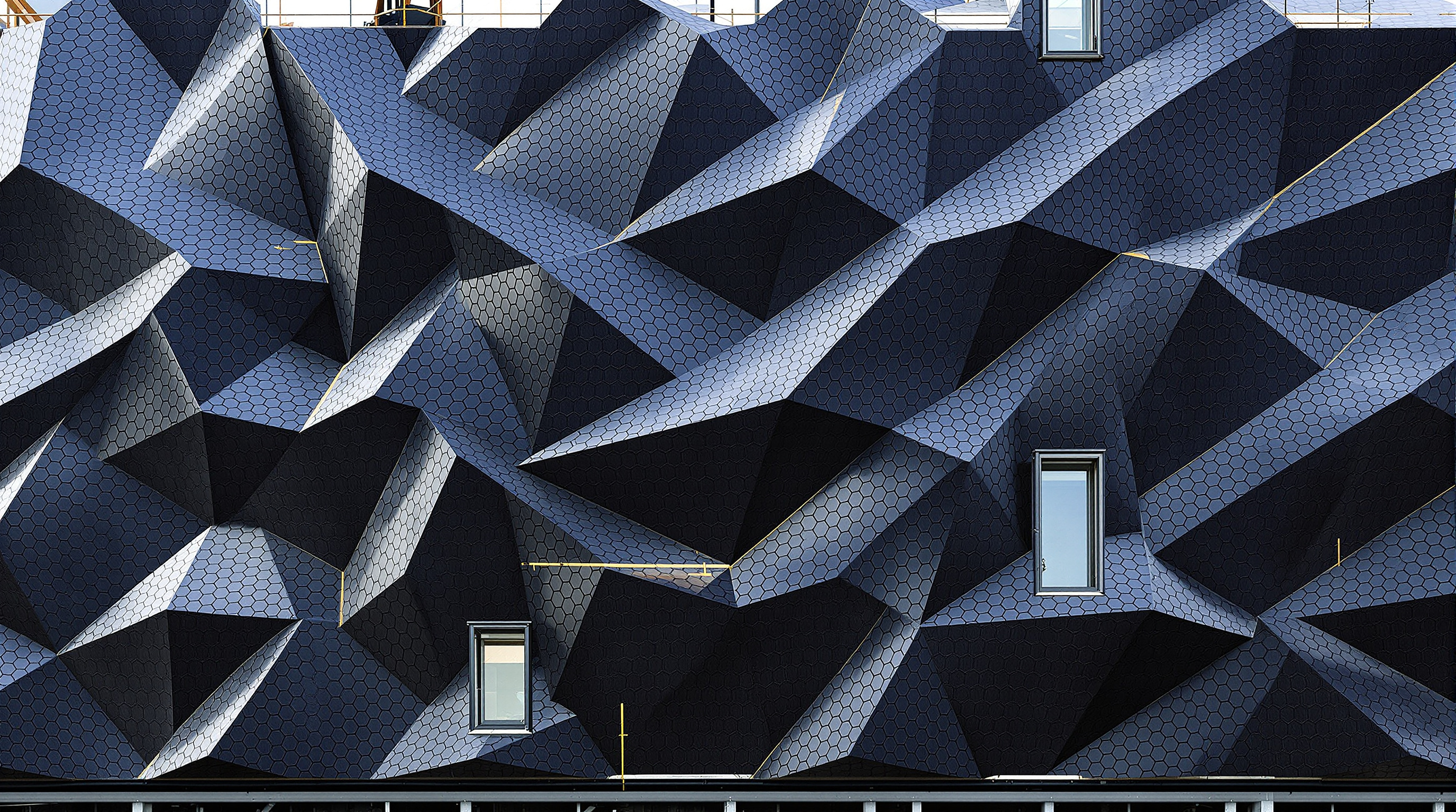 Close-up of Sun Rock's pleated facade under construction, dark shingled photovoltaic modules overlapping like fish scales across the folded surface, each facet tilted at a slightly different angle, with a few flush windows set into the array
