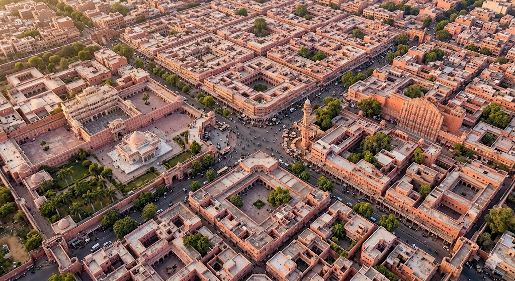 Wide-angle photograph of an urban design student sitting on the steps of a chaupar intersection plaza in Jaipur's old walled city in late afternoon, sketching the surrounding urban form into a hardcover sketchbook, the chaupar busy with the activity of the city — small shops opening to the plaza, people walking, a chai vendor with his cart, the pink facade language of Jaipur's regulated urban form visible all around, the elegant arched windows and jharokhas of the building facades providing a coherent ensemble, warm golden afternoon sun creating long shadows across the plaza, the student deeply absorbed in capturing the urban character through observation and drawing