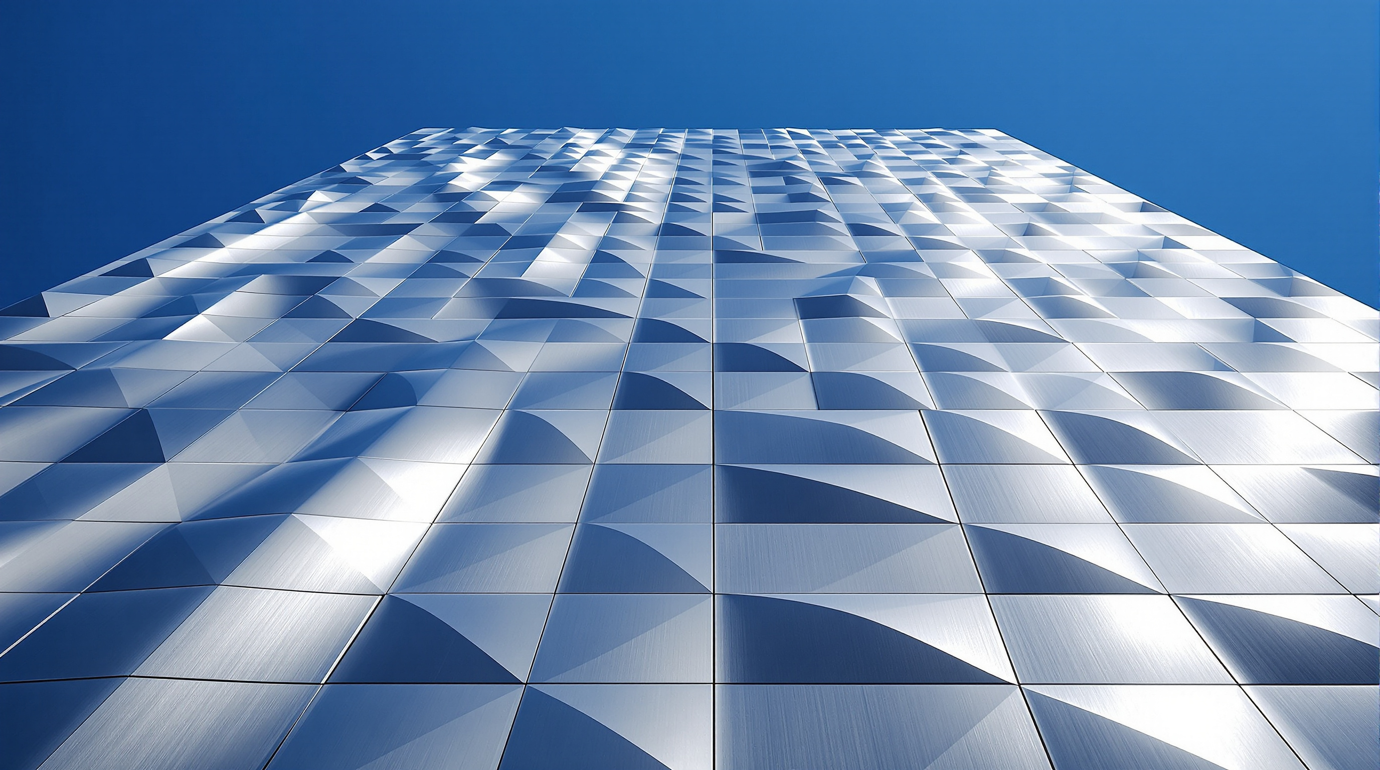 Looking up the great southwest face of VIA 57 West: a vast field of unique curved stainless-steel panels catching daylight, the wall visibly warping from vertical at the base to a steep inward slope, individual faceted panels reading as a rippling metallic skin against a blue sky