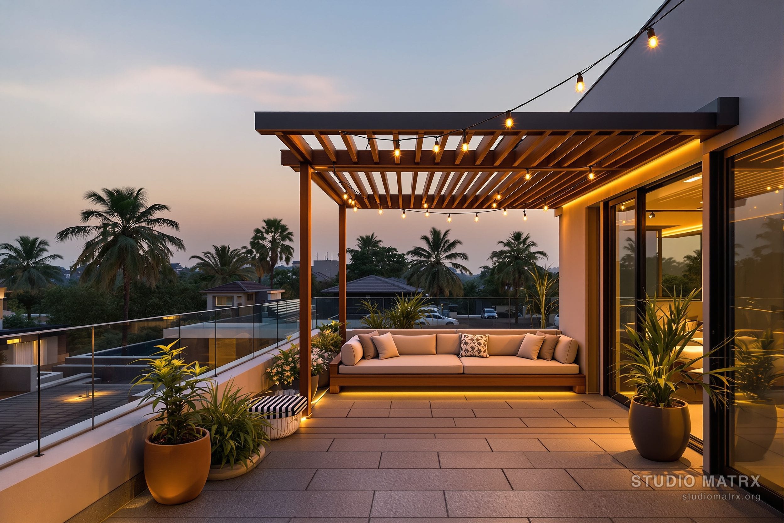 A private rooftop sky-deck on a modern South Indian home at golden hour — a timber pergola overhead casting striped shade, a built-in bench with cushions, planters of frangipani and ornamental grasses, a glass parapet, warm string lights, with palm trees and neighbouring rooftops beyond.