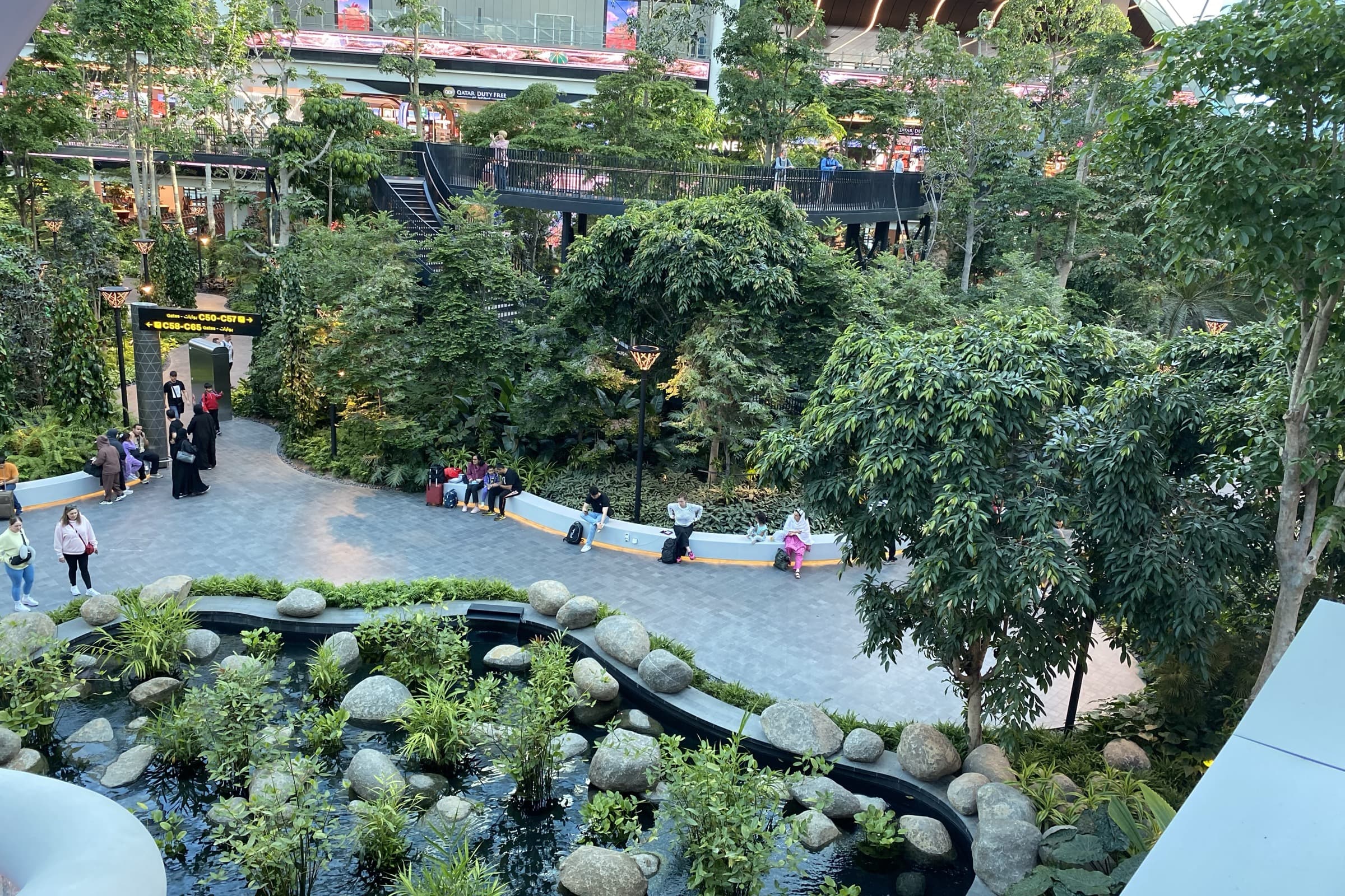 The Orchard at Hamad International Airport, Doha — a daylit indoor garden of tall trees with travellers resting beside a central water feature, terminal levels rising behind